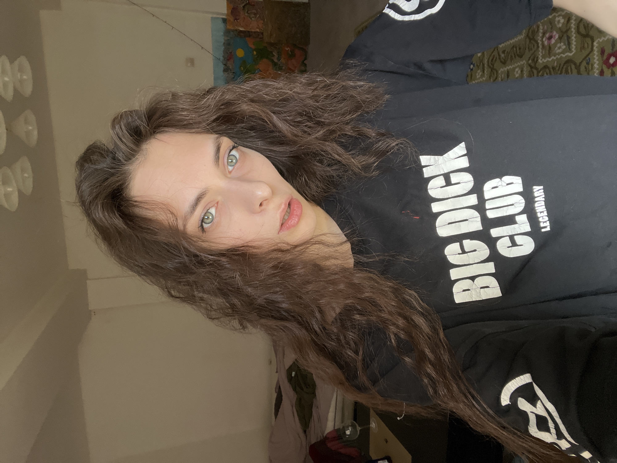 A person with long, wavy brown hair and light-colored eyes is wearing a black long-sleeved shirt with the words "BIG DICK CLUB" printed in white. The background includes a wall with a lighting fixture on the left, and what appears to be a rug and artwork on the wall in the upper-right. Below the person, items can be seen on the floor.