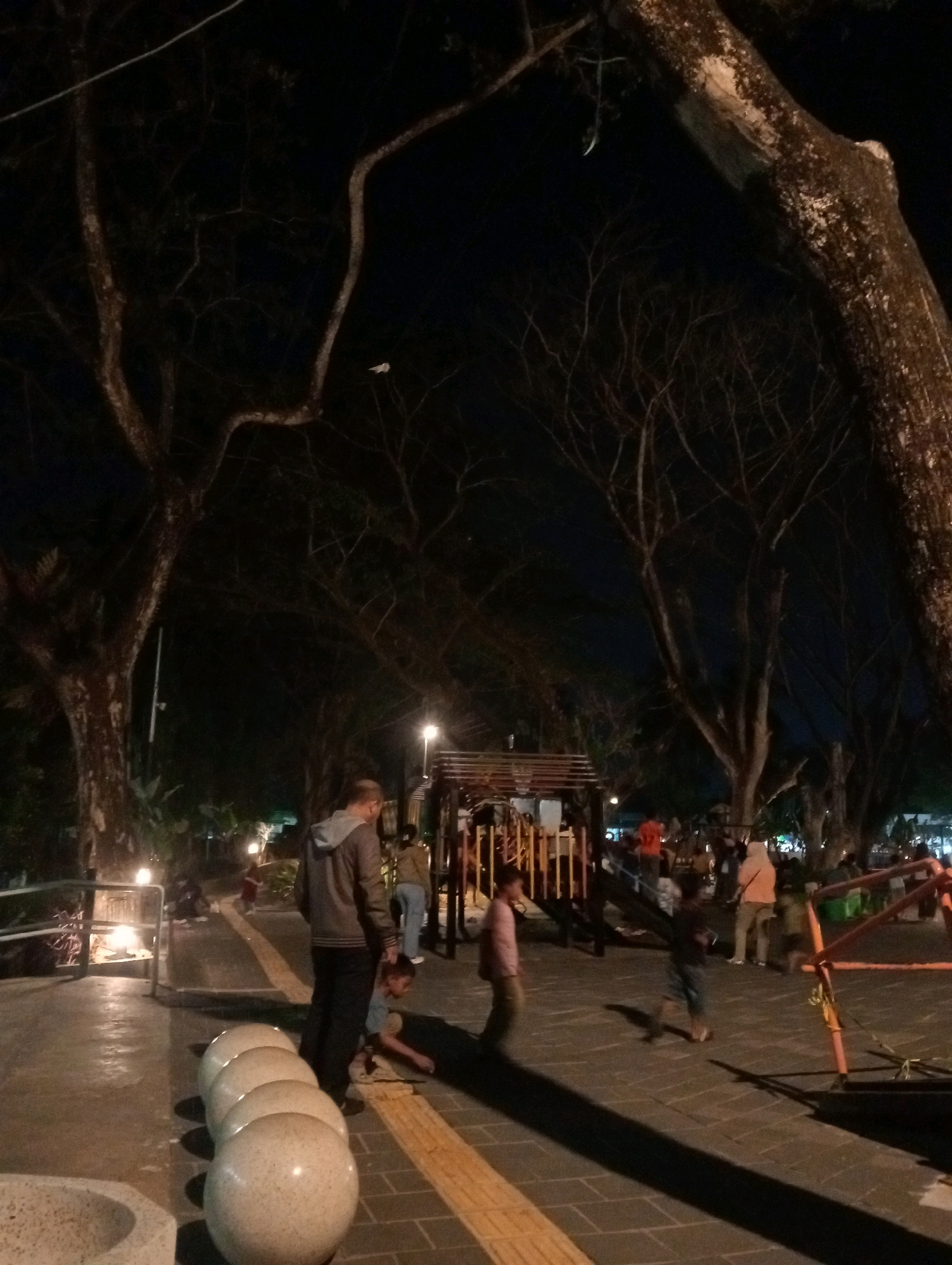 The image depicts a nighttime scene at a playground in Samarinda, Indonesia. The foreground features a row of large, rounded, light-colored stone objects, possibly seating or decorative elements, on a paved walkway with a textured yellow strip. To the right, a playground structure with orange metal bars is visible. Several individuals are present: a man in a gray hooded jacket stands facing away from the camera, his hands at his sides, while a child in a light-colored shirt is crouched near him. Further in the background, other people, including children and adults, are scattered around the playground, some interacting with the play equipment. The environment is characterized by large trees with bare branches silhouetted against a dark night sky. Artificial lighting from lamps illuminates parts of the scene, casting shadows and highlighting the textures of the pavement and the playground equipment.