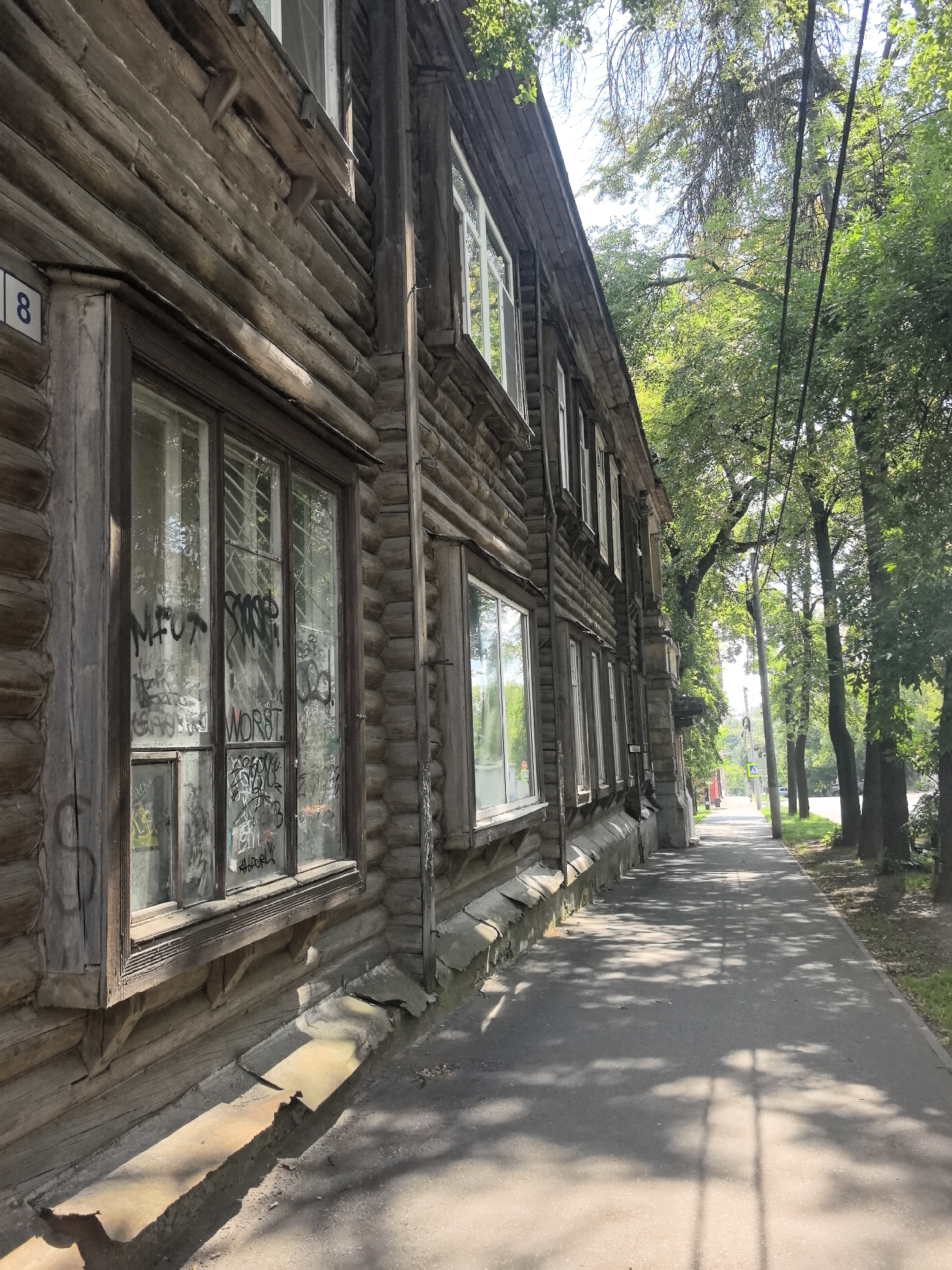 The image depicts a weathered wooden building, likely an apartment block, with visible graffiti on its windows. The building is situated in Nizhny Novgorod, Russia, along a paved sidewalk lined with trees. The sunlight casts shadows of the foliage onto the sidewalk, creating a dappled effect. The building itself appears to be constructed from logs, with multiple windows visible along its facade. The overall impression is one of an aging urban environment with a touch of urban decay. The specific location within Nizhny Novgorod is not identifiable from the image alone, but the architectural style and presence of trees suggest a residential area.