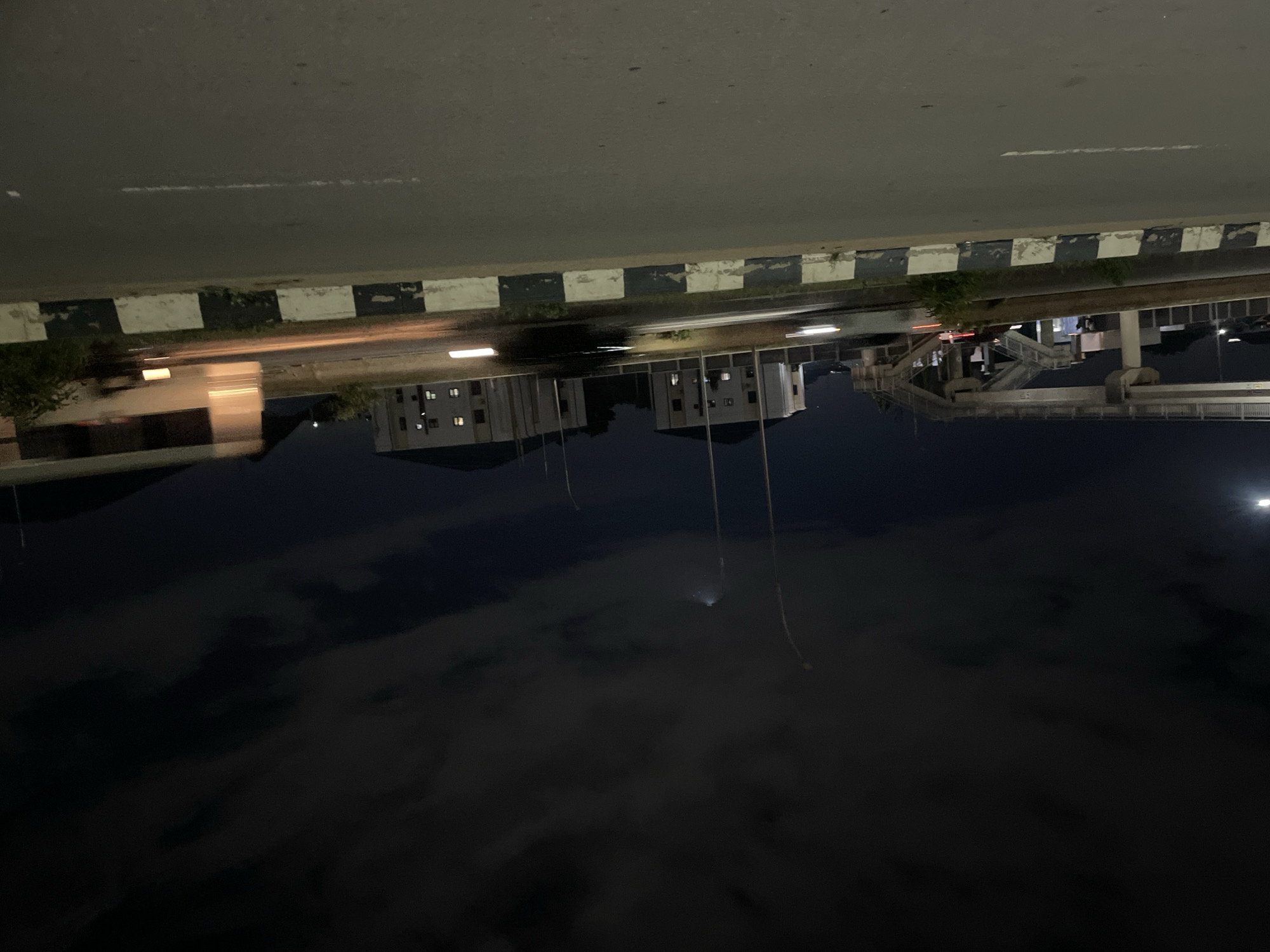 The image captures a nighttime scene on a road in Abuja, Nigeria, possibly an overpass area. The sky is dark with scattered clouds, illuminated by faint moonlight. Several streetlights are visible, casting a soft glow. In the background, modern multi-story buildings with illuminated windows stand against the dark sky. A pedestrian overpass, constructed with concrete pillars and a walkway, is prominent on the left side of the frame. The road itself is paved and shows markings. A vehicle, blurred due to motion, is visible on the road. A bus is also present, appearing as a streak of light and blurred form, indicating movement. A median strip with black and white markings separates the road lanes. The overall impression is of a well-lit urban environment at night.