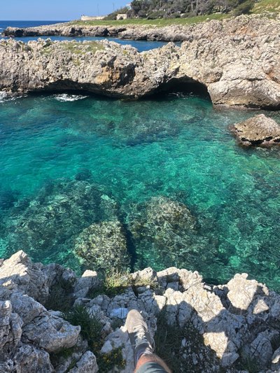 The image captures a coastal scene in Nardò, Italy, known for its rocky shores and clear waters. The focal point is a cove with vibrant turquoise water, revealing submerged rocks and the seabed. Rugged, light-colored limestone cliffs surround the cove, featuring small caves and sparse vegetation. In the foreground, the edge of the cliff is visible, with a person's foot and lower leg partially in the frame, suggesting the viewpoint is from someone standing on the rocks. In the distance, a building or structure can be seen amidst greenery on the elevated landmass, completing the coastal landscape.