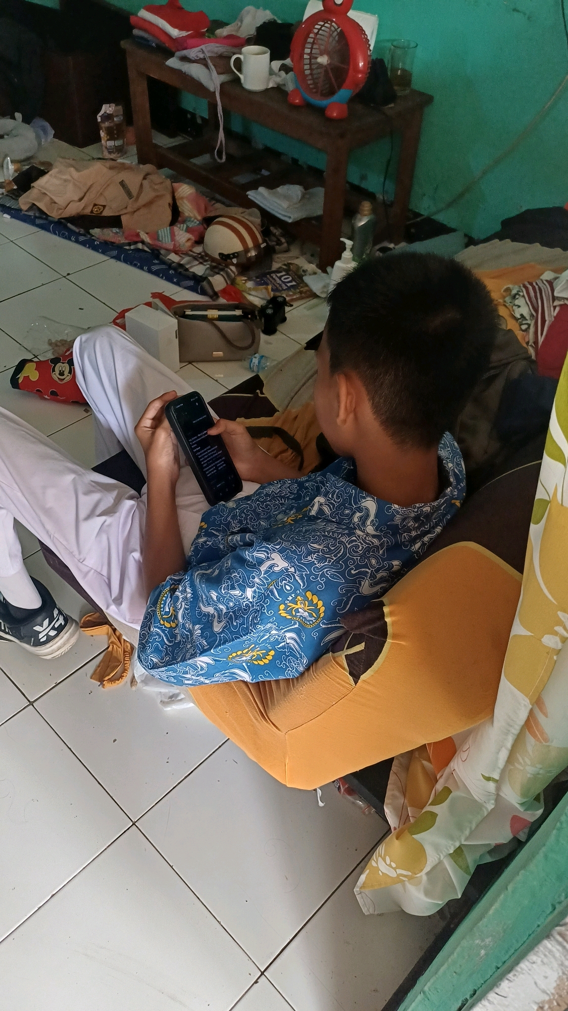 The image depicts a close-up, overhead view of a young person sitting on the floor, engrossed in their smartphone. They are wearing a white uniform shirt and pants, with a blue and white patterned garment draped over their shoulders. The setting appears to be a cluttered room with various items scattered around, including clothes, a helmet, a fan, a mug, and what looks like a magazine. The floor is tiled with a light-colored pattern. While the image itself does not provide explicit location markers, the style of the uniform and the general atmosphere suggest a domestic or casual educational environment within Indonesia, potentially in the city of South Jakarta. The focus is on the individual's interaction with their phone.