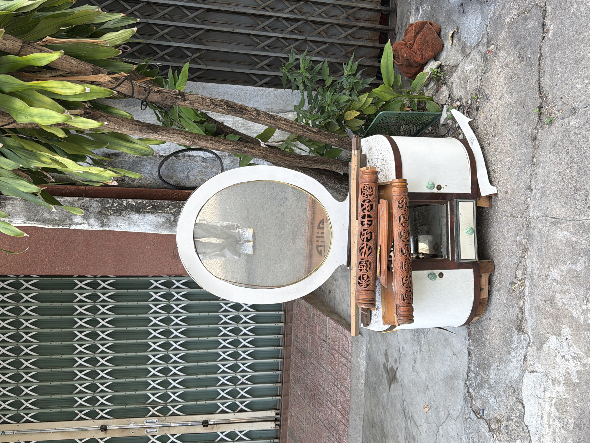 This is a street-level, eye-level, medium shot of a white and wood vanity with an oval mirror, placed on a concrete sidewalk in Nha Trang, Vietnam. The vanity is in disrepair, with visible damage to its wooden decorative elements and a missing side panel. A person's legs, wearing grey pants and white sneakers with yellow accents, are reflected in the mirror, suggesting the photographer is standing behind the vanity. The background features a closed, accordion-style security gate with a green metal lattice and a partial view of a building with a reddish-brown facade. To the right of the vanity, a tree with large green leaves grows, and further back, another accordion gate is visible. A crumpled brown fabric item and some debris are scattered on the ground next to the vanity. The lighting suggests it is daytime, with sunlight casting shadows. The scene appears to be an outdoor space in an urban setting, possibly a street or alleyway.
