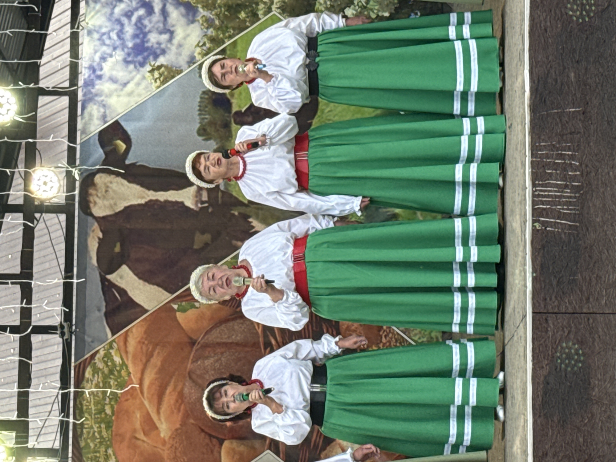 Four elderly women are standing on a stage in front of a backdrop that depicts a pastoral scene with a cow, bread, and sky. The women are wearing white blouses with ruffled collars and sleeves, red beaded necklaces, and green ankle-length skirts with white stripes near the hem. Each woman is holding a microphone and appears to be singing. They are wearing white headbands. The stage is set outdoors, indicated by the lighting and the backdrop. The city of Кукуи, Russia is mentioned as the location, but no specific landmarks or details within the city are visible in the image. The overall scene suggests a performance or cultural event.