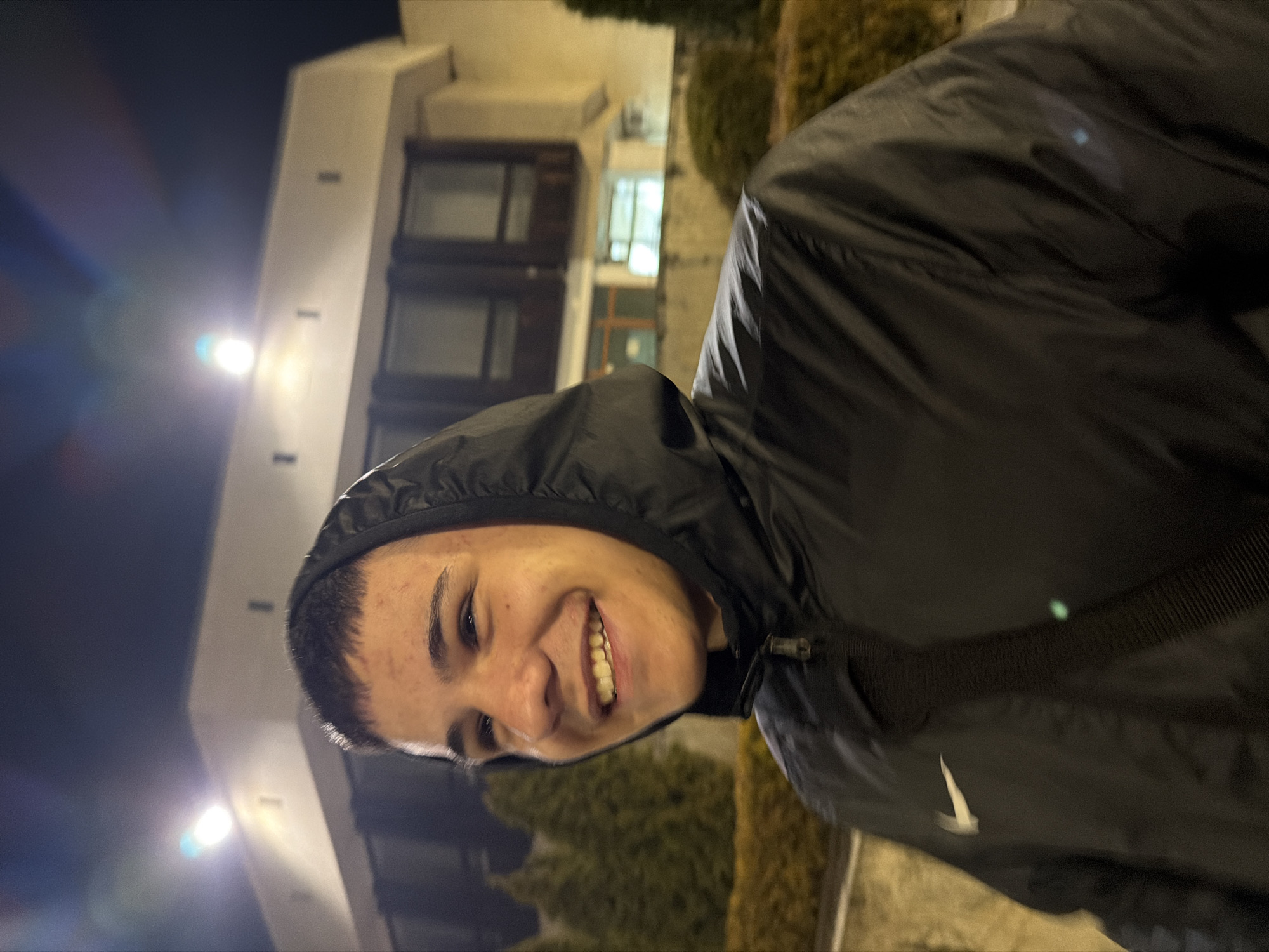 A young male with short hair and wearing a black hooded jacket with a white Nike logo is smiling towards the camera. He is positioned in front of a building with dark-colored double doors and illuminated by streetlights. Based on the building's style and location, it may be in Panagyurishte, Bulgaria. There are bushes nearby. The overall lighting suggests it is nighttime.