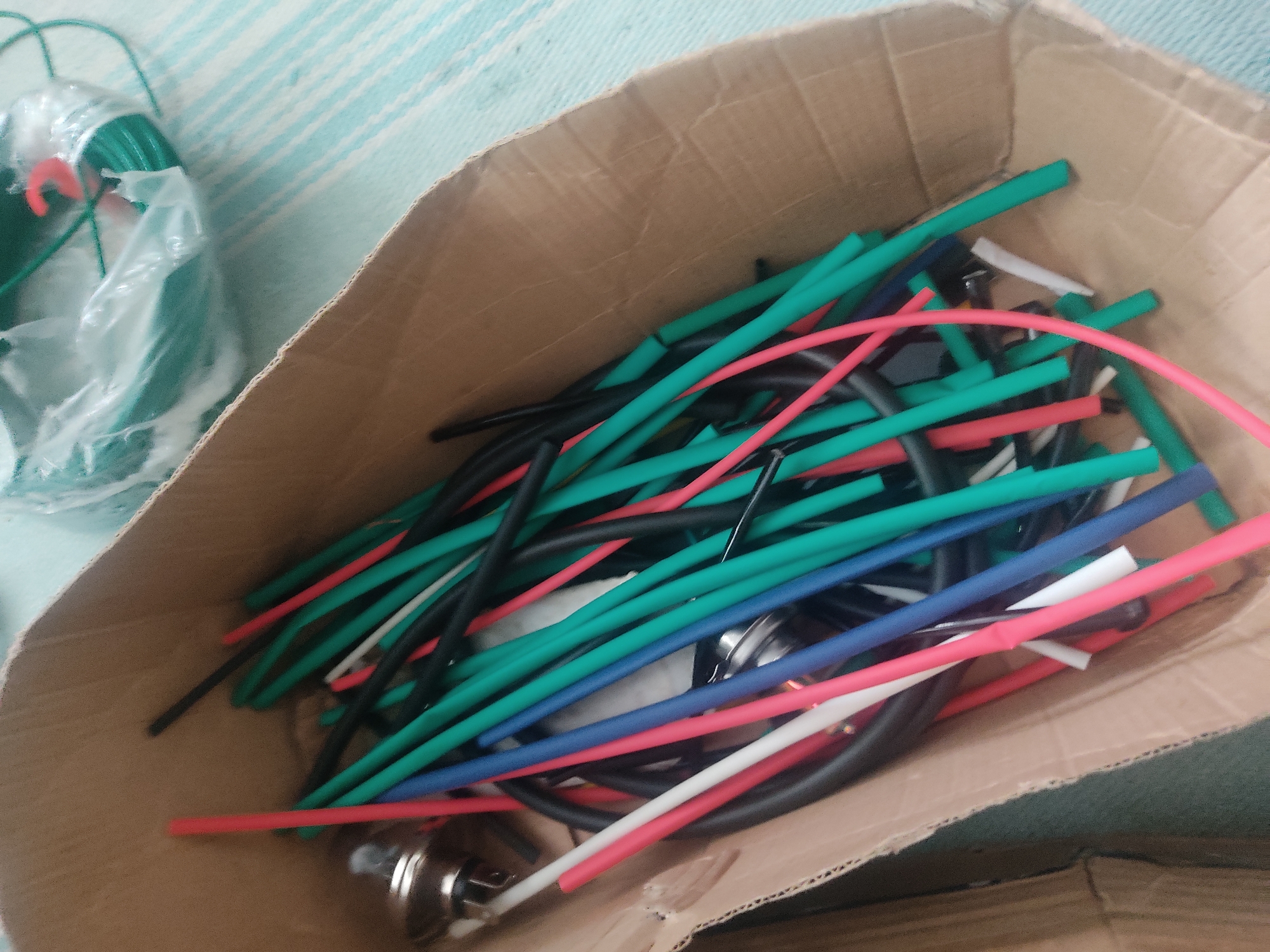A cardboard box is filled with various colored heat shrink tubing, appearing to be red, green, blue, black, and white. There are also a few metallic connectors visible amongst the tubing. In the upper right corner, a roll of green wire is partially visible, wrapped in plastic. The box is situated on a striped surface, likely a rug or tablecloth, with pale blue and white stripes. The overall scene appears to be indoors, with no specific city or location details discernible.
