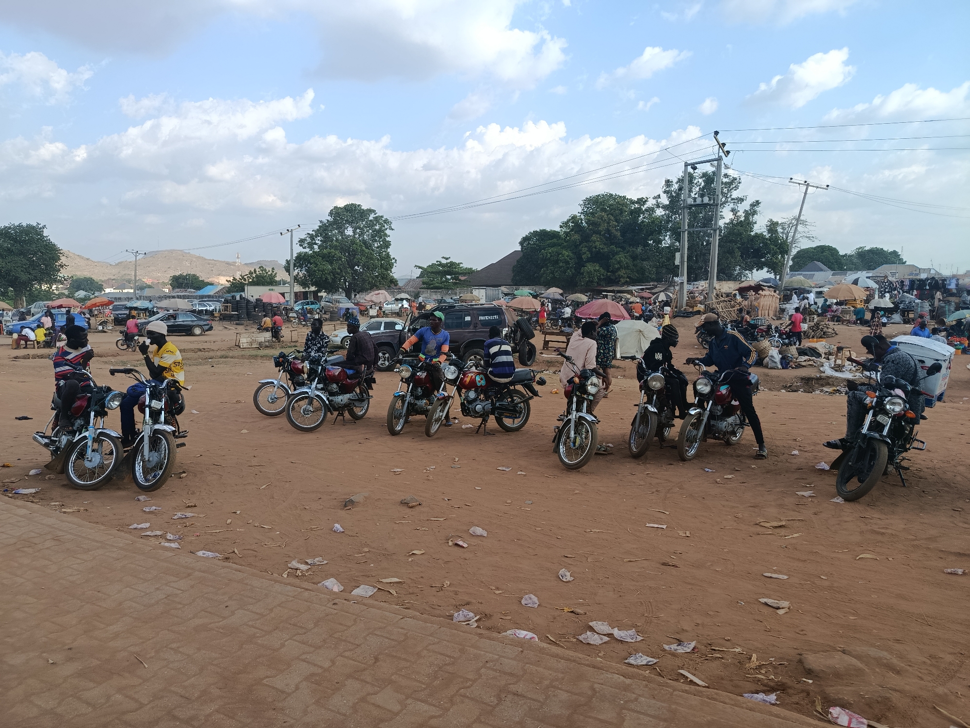 The image depicts an outdoor scene at a marketplace in Gbessa, Nigeria. Several motorcycles are parked on a dirt lot littered with debris and plastic waste. The marketplace is crowded with people and stalls covered with umbrellas, indicating potential commercial activity. A mix of vehicles including cars and motorcycles, are visible within the market area. Overhead power lines are visible. The landscape includes trees, distant hills, and a sky with clouds. The foreground shows a paved area adjacent to the dirt lot. Individuals are seen standing or sitting on the motorcycles.