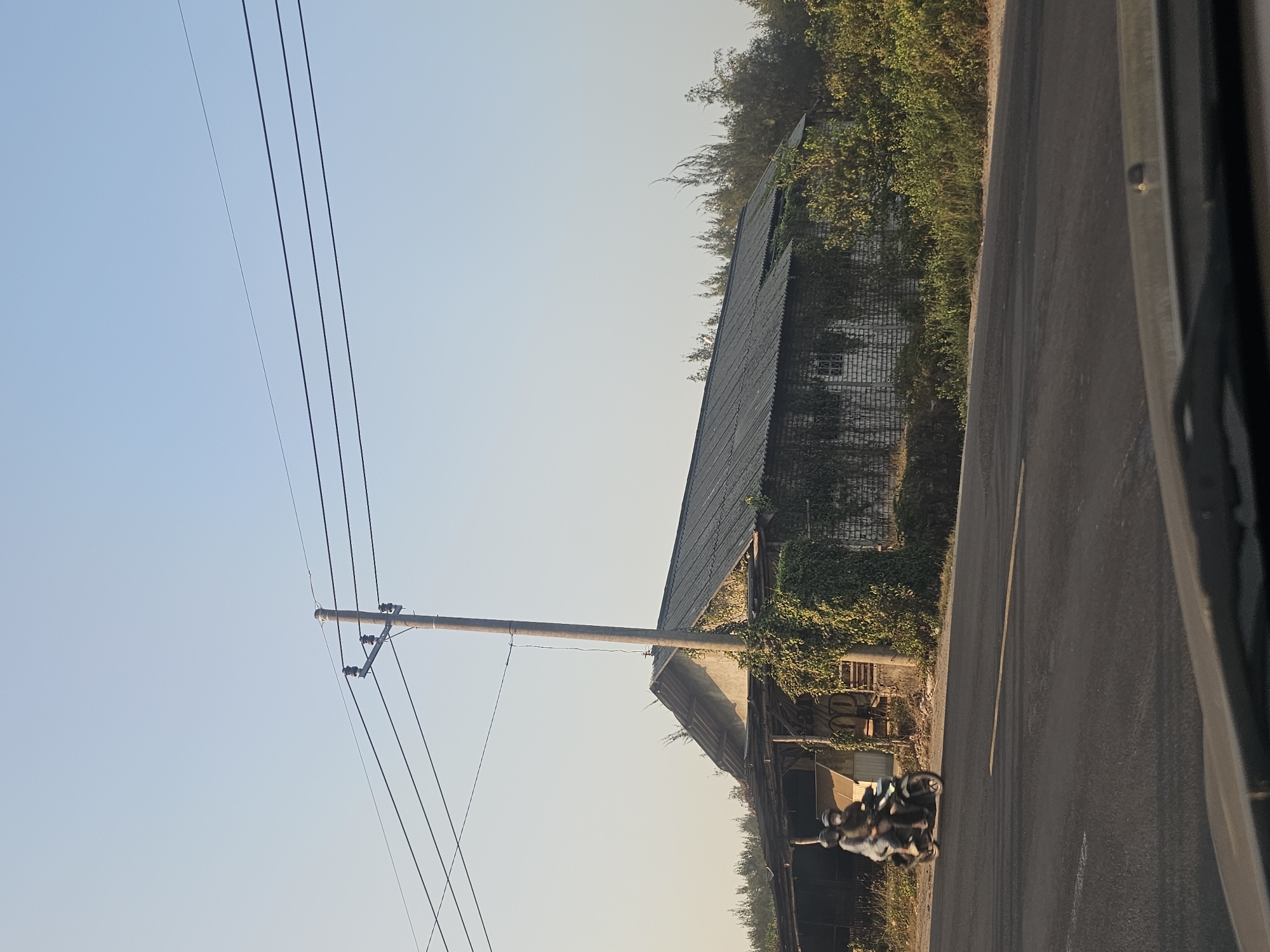 The image depicts a rural or semi-rural scene in Jenu, Indonesia, with a focus on a long, low-slung building with a corrugated metal roof. The building appears aged, with white brick walls partially obscured by thick green vines and ivy. Several windows are visible, some appearing as dark openings and others with a criss-cross pattern. The structure has an open-sided section on the left, resembling a covered patio or storage area. A tall utility pole with multiple wires runs vertically in front of the building, casting a shadow.
In the foreground, a paved road with a yellow line is visible. A motorcycle with two occupants, both wearing helmets and masks, is traveling on the road from left to right. The occupants appear to be wearing light-colored clothing. Behind the building, dense green foliage and trees are present. The sky is clear and a pale blue, suggesting it is daytime, possibly late afternoon given the lighting and gentle haze. The overall impression is one of a somewhat disheveled but functional property in a natural setting.