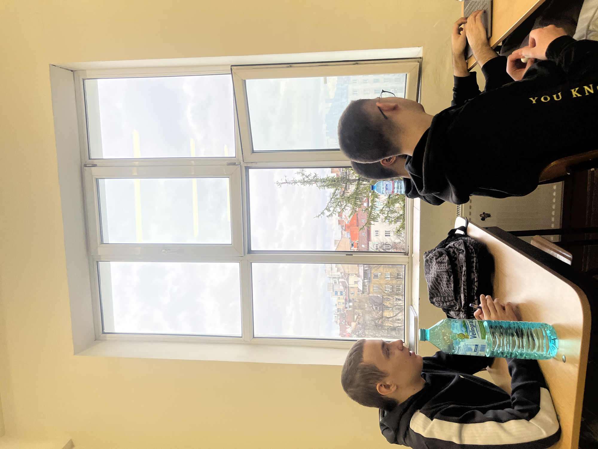 Two male individuals are present in the image, seated at adjacent desks. One man is wearing glasses and a black hoodie with the words "YOU KN" visible. He is working on a laptop. The other individual is wearing a black hoodie with white stripes. They are sitting beside a window. On the second man's desk is a blue water bottle and a pen. A backpack is on the floor between the two desks. Through the window, an urban landscape is visible. The buildings, with orange and red rooftops, and the leafless trees suggest the city is Sofia, Bulgaria.