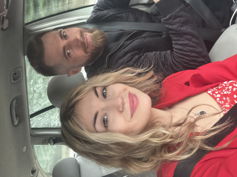 Two individuals are seated inside a vehicle, occupying the front passenger and driver's seats, with both wearing fastened seatbelts. The camera perspective is from within the vehicle, capturing a selfie-style image.
In the foreground, a light-skinned female with wavy, shoulder-length blonde hair with darker roots is positioned to the left. She is smiling, looking directly at the camera. She wears a red blazer over a white garment with a red floral pattern. A dark, oval-shaped pendant hangs from a silver chain around her neck.
Behind and to the right of the female, a light-skinned male is visible. He has short dark hair and a neatly trimmed beard. He is also smiling and looking towards the camera. He wears a dark, possibly black, quilted jacket. His left arm is visible, resting near the steering wheel (not fully visible).
The interior of the vehicle features light gray upholstered seats and headrests. Above, grab handles and interior light fixtures are partially visible on the ceiling. Through the side and rear windows, blurred green foliage is visible, indicating an outdoor setting, potentially a roadside or natural landscape.
Based on visual evidence, the specific location within the city of Budva, Montenegro, cannot be determined from the image.
