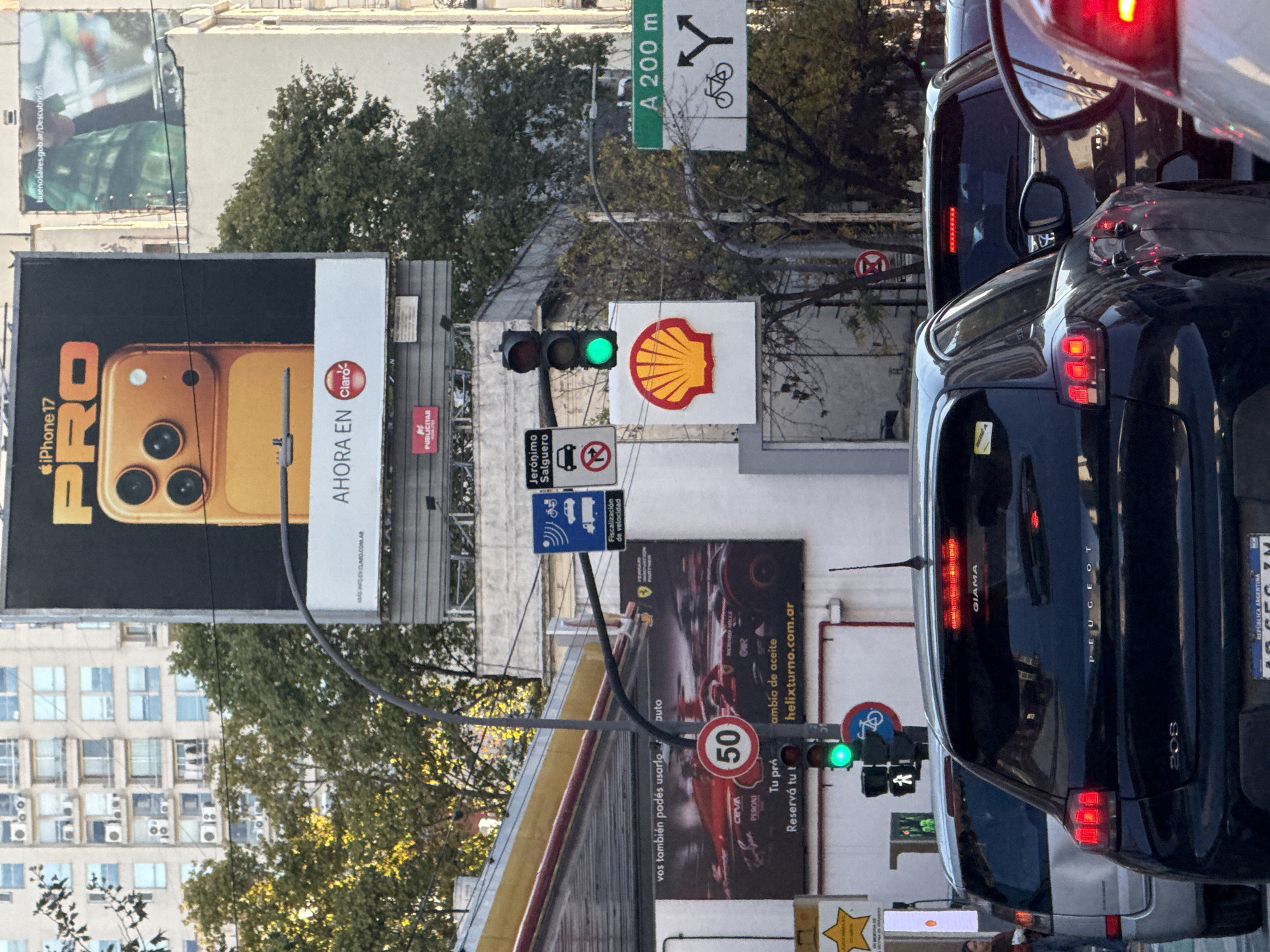The image captures a vibrant urban street scene in Buenos Aires, Argentina, under bright daylight. In the foreground, a dark grey/black Peugeot 208 hatchback is prominently featured from the rear, its taillights visible, with "208" and "GIAMA" badges, and "REPUBLICA ARGENTINA" on its license plate. An SUV or van is stopped in front of it, reflecting the halted traffic at what appears to be a green traffic light.
The street is bustling with advertisements and road infrastructure. A large billboard on the left displays a gold "iPhone 17 PRO" with "AHORA EN Claro" and "SINFDEN CLARO.COM.AR". Below it, another sign reads "buenosaires.gob.ar/DescubrirBA". To the right, a prominent Shell logo sign is visible, alongside a speed limit sign indicating "50". Another billboard features a red Formula 1 car, advertising "ambio de aceite" (oil change) at "helixturno.com.ar", with additional text like "vos también podés usarlo," "CEVA," "PERONI," "Tu pró," and "Reservá tu t."
Various road signs include a green directional sign pointing "A 200 m" for a split road with a bicycle lane, a blue sign indicating "Fiscalización de velocidad" (speed enforcement) with vehicle and radar symbols, and a sign identifying the street as "Jerónimo Salguero" with a 'no U-turn' symbol. The background shows multi-story buildings with numerous windows and air conditioning units, interspersed with green trees. The scene depicts a typical busy city environment with commercial activity and vehicular traffic.