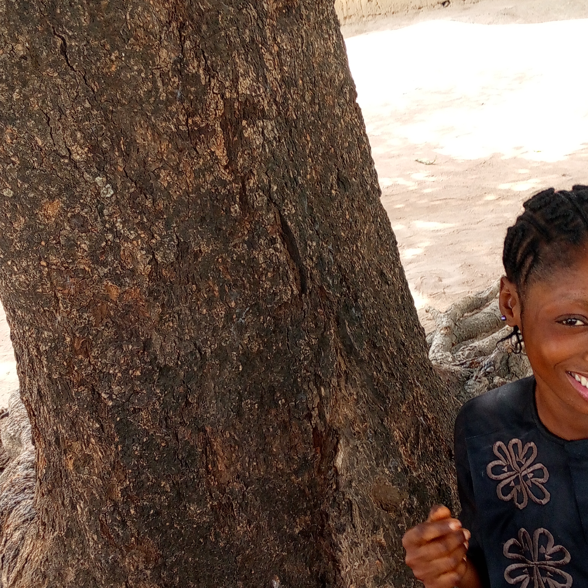 The image shows a smiling woman standing beside a large, dark-barked tree. She is wearing a dark shirt with floral embroidery. The background appears to be a sandy or dirt surface, with hints of a building in the distance. The location is identified as Mararaba, Nigeria, though no specific landmarks or features identifying the city are clearly visible. The primary subjects are the woman and the tree. The main action is the woman smiling, which suggests a positive interaction with the environment or the photographer.