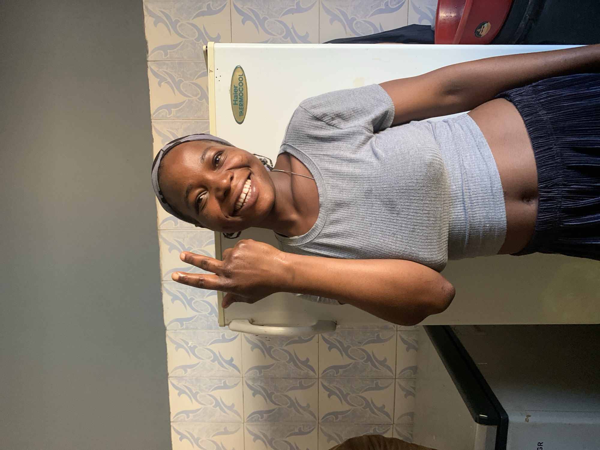 A young woman with a bright smile and a grey headband is posing in what appears to be a kitchen in Jos, Nigeria. She is wearing a grey, ribbed crop top and dark, pleated pants. Her left arm is raised, and she is holding up two fingers in a peace sign. Her right hand is not fully visible. Behind her, a white Haier Thermocool refrigerator stands against a wall tiled with a light blue and white patterned design. To her left, a dark countertop with some equipment is partially visible. The overall lighting suggests it's daytime, possibly with sunlight coming from the side, casting shadows.