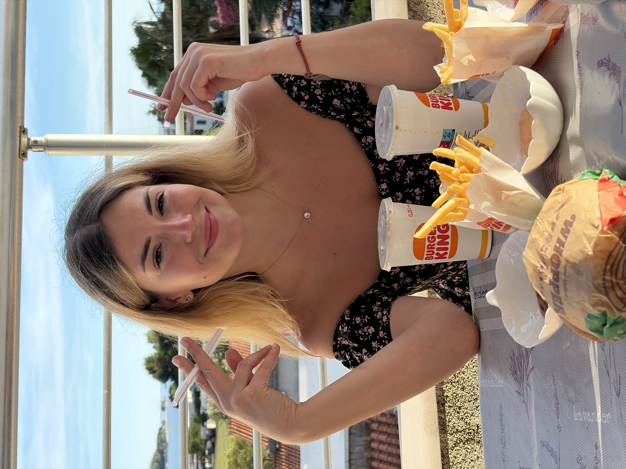 The image shows a young Caucasian woman with blonde hair, wearing a black floral print dress, sitting at a table outdoors. She is holding two drinking straws, one in each hand, in a way that resembles smoking. In front of her are two Burger King cups and two servings of french fries in paper containers. A hamburger wrapper is also visible on the table. The background reveals a view of the Adriatic Sea and buildings with red tile roofs, characteristic of Budva, Montenegro. The woman is smiling and looking at the camera.