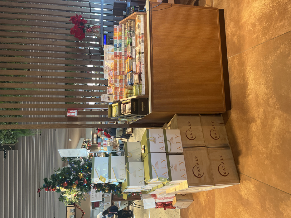 This indoor shot, oriented vertically, captures a bustling commercial space, likely a specialty food market or store, decorated for the holiday season. In the mid-ground, a long, light wooden display counter stretches across the frame, laden with numerous packaged food items, including various candies, chocolates, and what appear to be gourmet snacks, meticulously arranged in rows. Below and to the right of this counter, several light wooden crates are stacked, prominently featuring the red "EATALY" logo on their sides, suggesting an Italian food retail outlet.
To the left of these crates, a collection of elegant gift bags, primarily in white with gold and green accents, are stacked high. The letters "ALY" are visible on some of these bags, possibly part of a larger brand name. Beneath these bags, red and white gift-wrapped boxes are also visible, contributing to the festive atmosphere.
In the background, a fully decorated Christmas tree, adorned with colorful ornaments and lights, stands prominently to the left, further emphasizing the holiday setting. Behind the tree and display counter, a slatted wooden partition or wall creates a textured backdrop. A red sign, likely indicating a fire extinguisher, is mounted on this partition. A few people can be faintly seen in the deeper background, with one person wearing a red Santa hat. The floor is covered in large, textured reddish-brown tiles. The overall scene suggests a vibrant retail environment, possibly in Dorno, Italy, given the Eataly branding and the context provided.