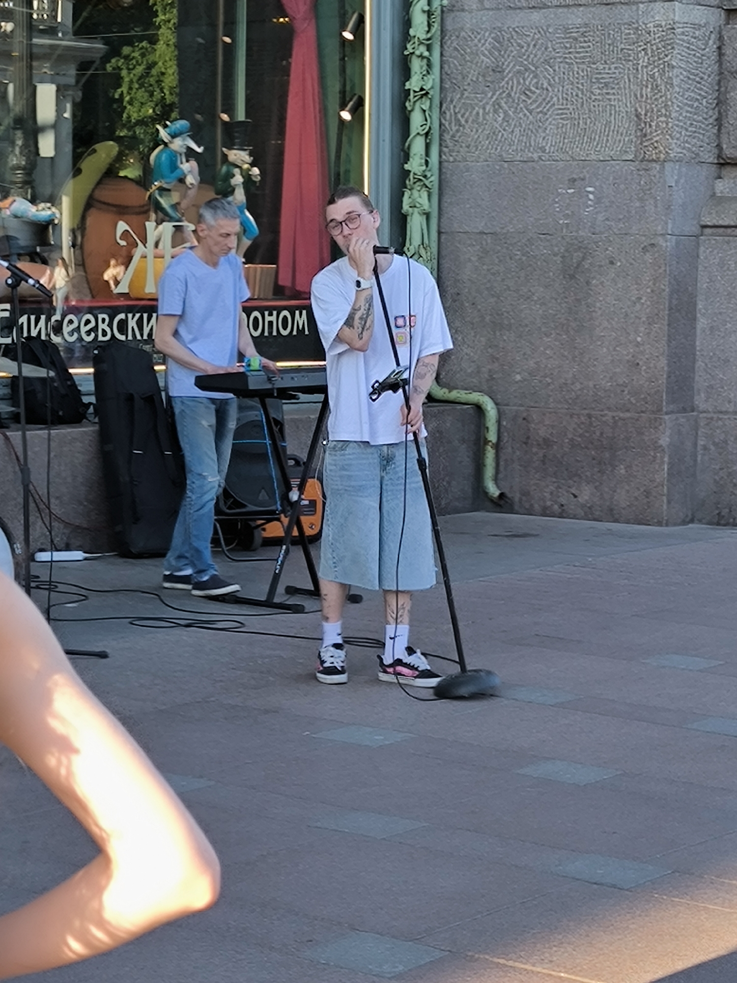 The image shows two male street musicians performing or preparing to perform on a paved sidewalk in front of the Eliseevsky Gastronom at Nevsky Prospekt, Saint Petersburg, Russia, on a sunny day.
The man on the left has short, grey hair and wears a light blue t-shirt and light blue distressed jeans with dark sneakers. He is positioned behind a black electronic keyboard on a stand, with his hands over the keys. To his left, a black equipment bag and a speaker are on the ground, with various cables visible.
The man on the right is younger, wearing eyeglasses, a white t-shirt, and light blue denim culottes, with white socks and dark sneakers featuring pink accents. He has visible tattoos on both arms and legs. He stands in front of a black microphone on a stand, with his left hand near his mouth. A green drainpipe is visible on the grey stone building wall behind him.
The background features the ornate exterior of the Eliseevsky Gastronom building, characterized by decorative green columns and a textured grey stone facade. Behind the keyboard player, a large display window reveals theatrical-looking figures, including one in blue and another in green with a top hat, a large brown barrel, and part of a yellow sign with Cyrillic text, including "Ж" and "ЕЛИСЕЕВСКИЙ" (Eliseevsky) and "ОНОМ" (part of Gastronom). A red curtain is visible inside the shop window.
The ground is paved with rectangular light grey and darker grey tiles, and strong sunlight casts shadows, highlighting a partially visible arm in the lower left foreground.