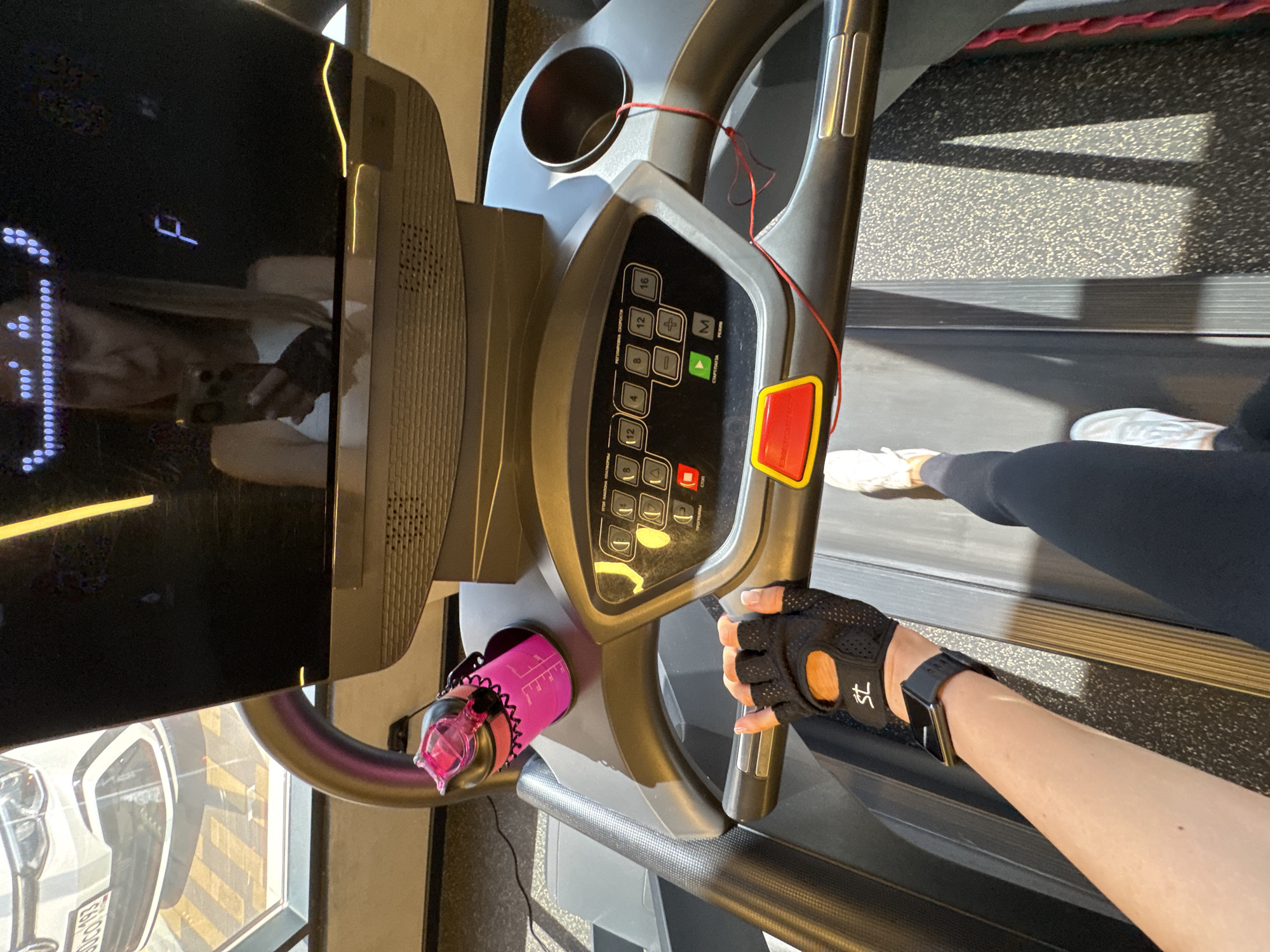 The image shows a first-person perspective of a person using a treadmill in a gym. The person is wearing black workout gloves and a smartwatch. They are running on the treadmill, as indicated by their legs and feet in motion. A pink water bottle is clipped to the side of the treadmill. In the background, a large screen displays a reflection of a woman holding a phone and a letter "F". A window behind the treadmill shows a car and a yellow striped pattern. The gym environment appears modern, with a black textured floor and bright lighting. The presence of a car with a Russian license plate (GCG 193) visible through the window suggests the location could be Sochi, Russia.