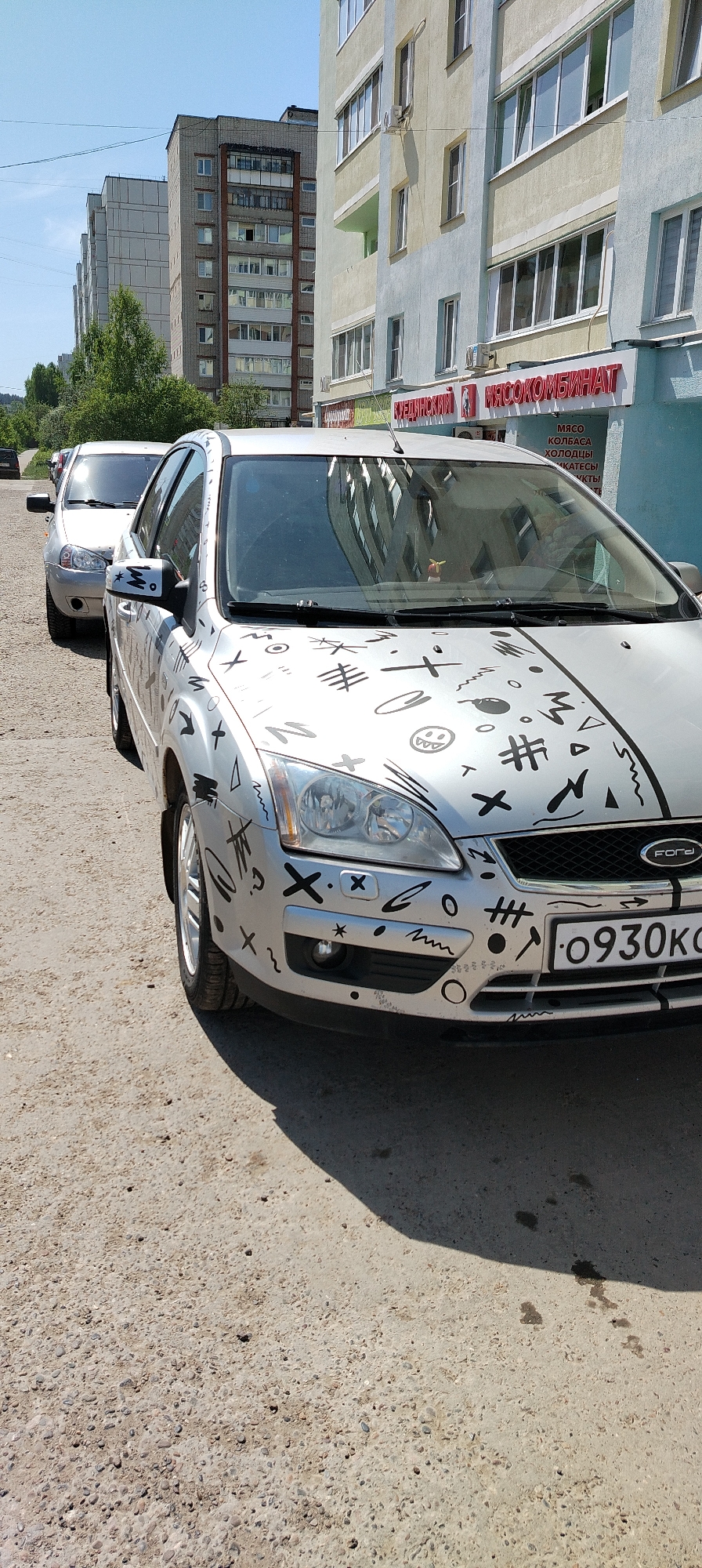 The image shows a silver Ford Focus car parked on a cracked concrete surface in Chaikovsky, Russia. The car has black graffiti-like markings on its hood and front bumper. The license plate reads "O930KC". Behind the Ford, another silver car is partially visible. A multi-story building is in the background. A sign above the ground floor says, in Russian, “Meat Processing Plant.”