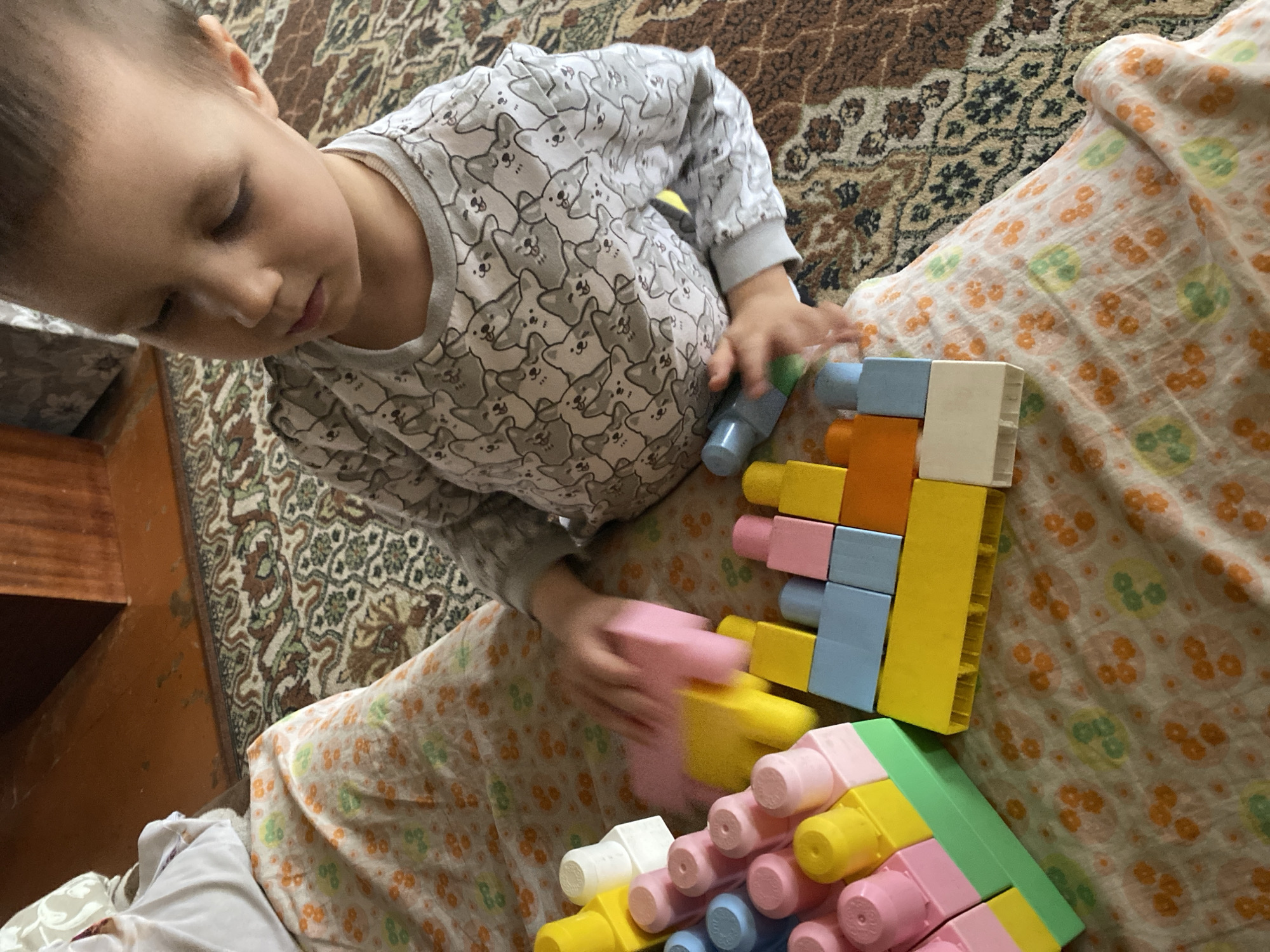 A child is playing with colorful building blocks on a bed with a patterned blanket. The child wears a long-sleeved shirt with a pattern of dog-like creatures. The setting appears to be indoors, possibly in a home. The location is unknown.