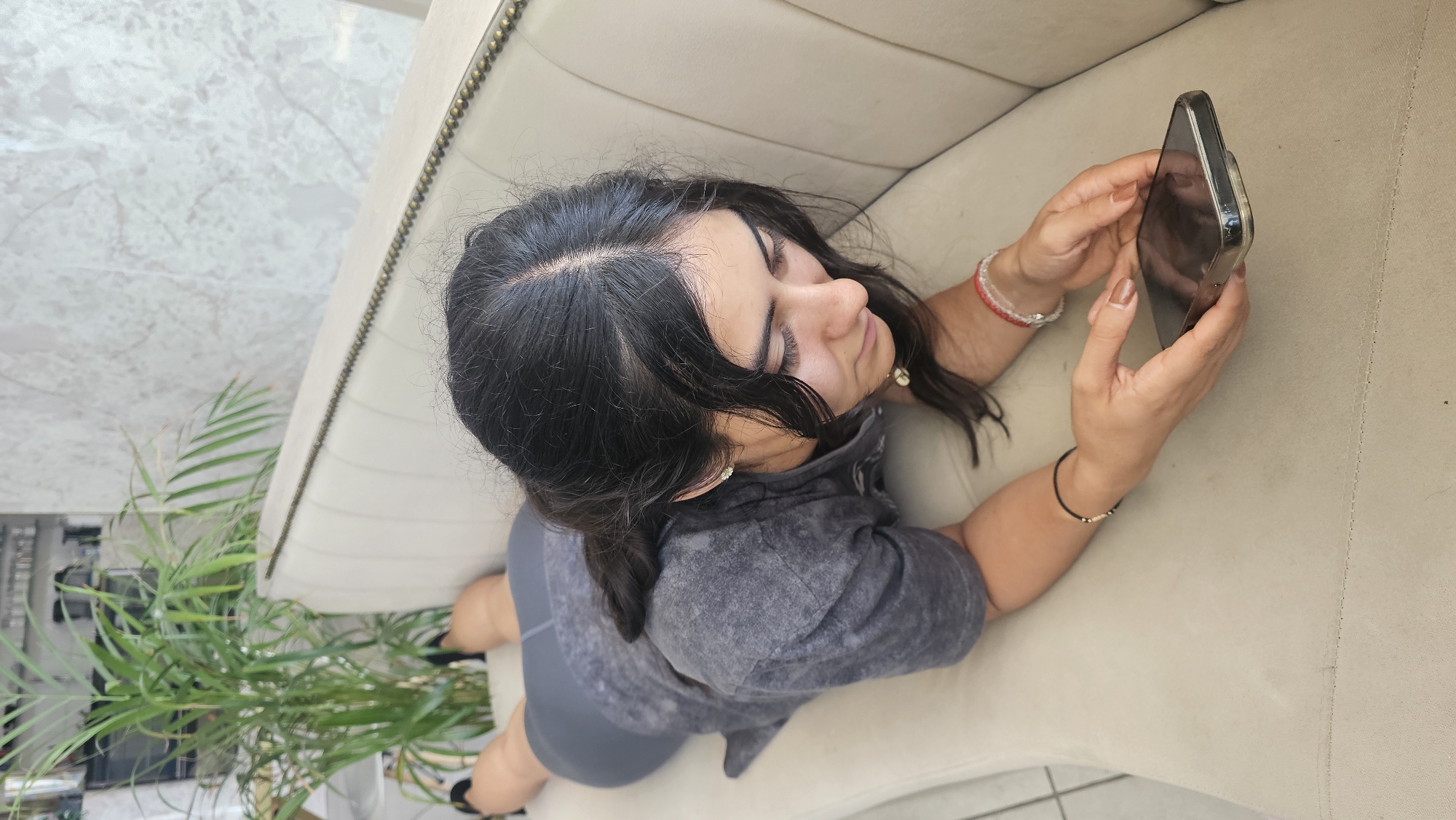An overhead shot shows a young woman with dark hair, dressed in a gray short-sleeved top and matching shorts, sitting on a light beige couch. She is engrossed in her smartphone, holding it with both hands and gazing at the screen with a focused, almost serene expression, her eyes appearing nearly closed. Her nails are painted a neutral brown color, and she wears bracelets on both wrists – a red and white beaded one on her right, and a black string with small beads on her left.
To her left, a vibrant green potted plant with long, slender leaves adds a touch of nature to the indoor setting. The couch features decorative brass studs along its top edge, suggesting a classic or ornate style. Behind her, a light-colored wall with a subtle, swirling marble-like pattern is visible, while the floor appears to be tiled. The overall lighting suggests it is daytime. The scene depicts a moment of quiet concentration or relaxation, taking place in Puebla, Mexico. No visible text is discernible in the image.