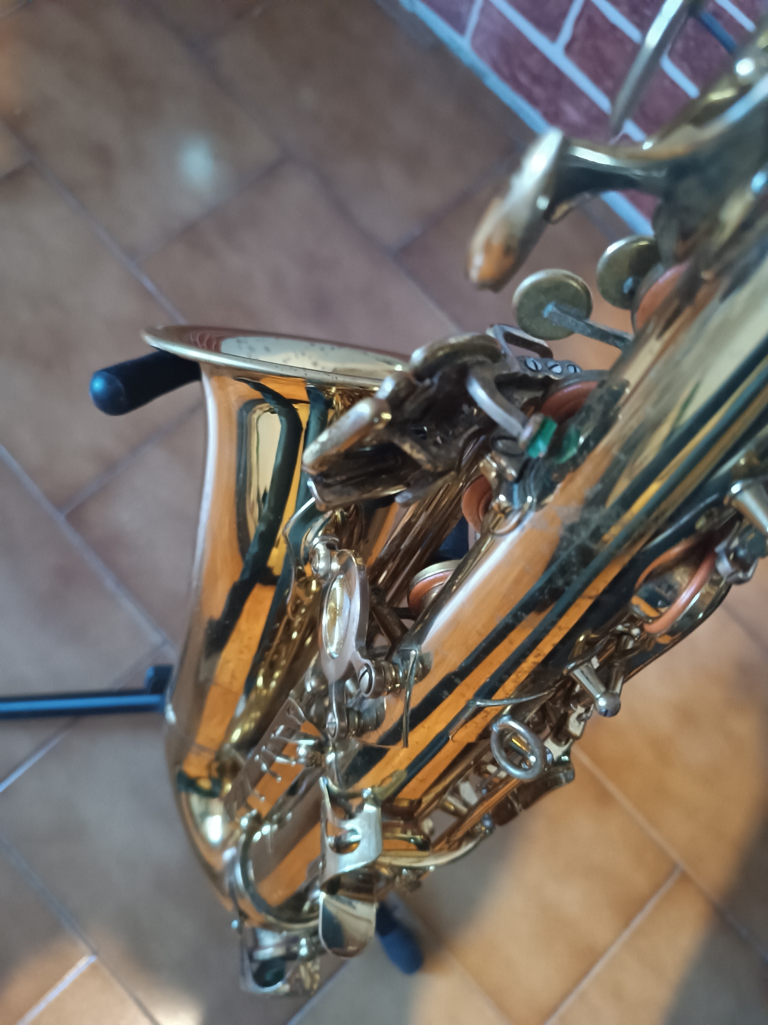 The image shows a close-up view of a saxophone standing on a tiled floor. Parts of a brick wall are visible in the background. The lighting creates strong reflections on the brass surface of the instrument. There are no people visible in the scene. The setting might be indoors, potentially in Viadana, Italy, given the instruction.