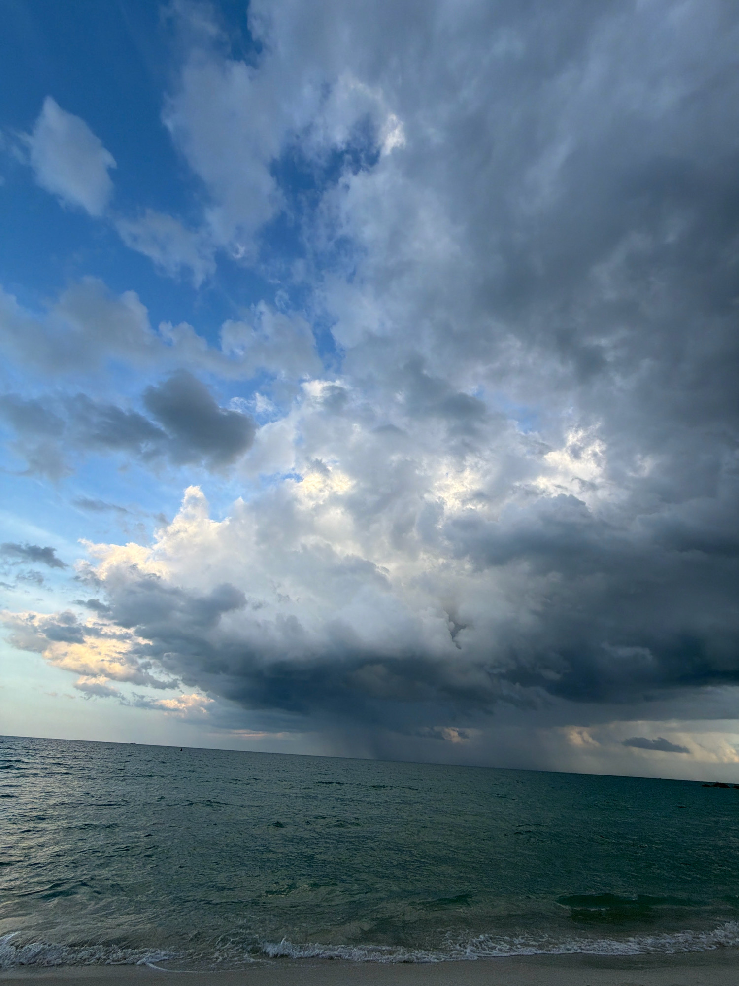 The image shows a stormy sky over the ocean. Dark, heavy clouds dominate the upper portion of the frame, with streaks of rain visible in the distance. Patches of lighter sky and clouds are visible breaking through the storm. The ocean is a deep teal color, with moderate waves reaching the sandy shore in the foreground. The scene appears to be a beach in the Ko Samui District of Thailand, as suggested by the tropical vegetation visible on the distant horizon. There are no people or specific objects visible in the image, making it a purely environmental scene.