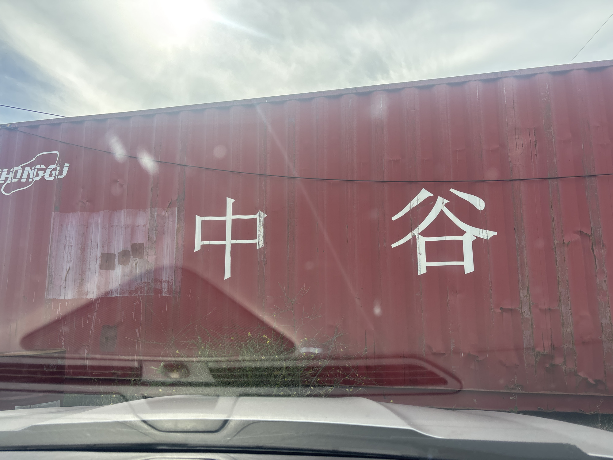 The image shows a close-up of a large, red shipping container with white Chinese characters painted on its side. The characters appear to be "中" and "谷". To the left of the Chinese characters, there is a white logo that reads "ZHONGGU" in a stylized font. The shipping container has visible signs of wear and tear, with some rust and discoloration. The container is partially obscured by a reflective surface, likely a car windshield, in the foreground. Through the windshield, a glimpse of weeds and small yellow flowers can be seen growing at the base of the container. The sky above is partly cloudy with sunlight streaming through, creating lens flares and reflections on the container's surface. The scene is likely an industrial area or a transportation hub in the city of Tbilisi, Georgia, given the presence of shipping containers and the overall environment.