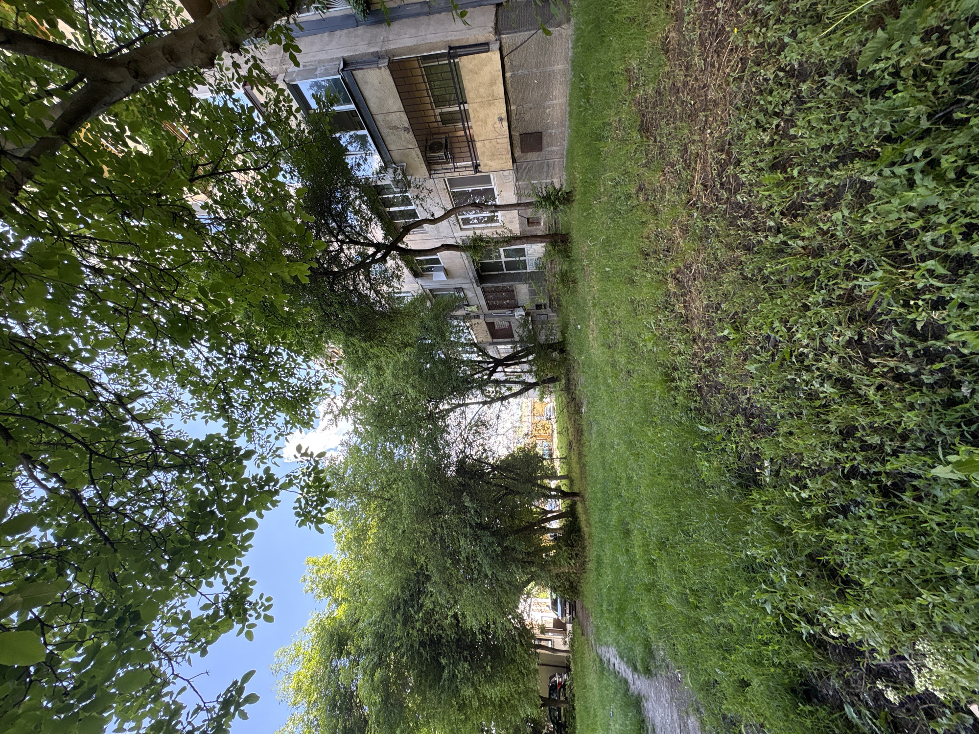 A building is positioned to the left of a grassy walkway and is partially obscured by lush green trees. The image shows a ground-level view, suggesting a street scene or a park setting. The architectural style of the building features elements common in urban environments, possibly within the city of Sofia, Bulgaria, given the information provided. No people are visible.