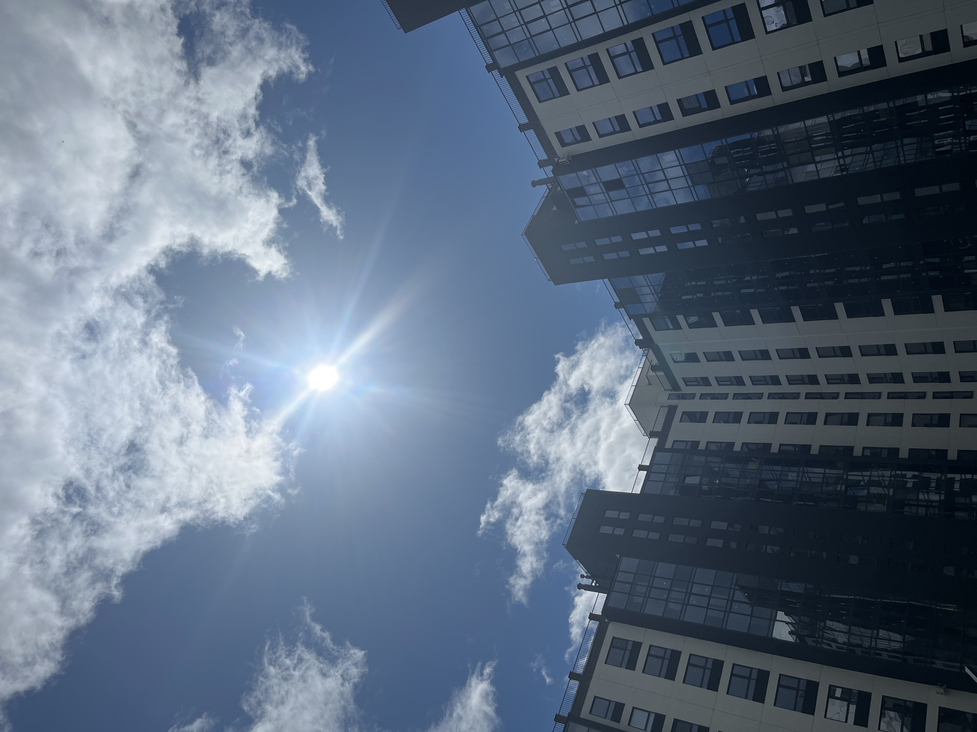 A low-angle, outdoor, vertical shot captures a modern apartment building under a bright, partly cloudy blue sky with the sun shining brightly. The building's facade is a contrast of light-colored panels and dark, reflective glass windows arranged in a grid pattern. Some sections of the building are darker, appearing to be more heavily glazed. The top of the building features black railings along balconies. Wispy white clouds are scattered across the sky, with the sun's rays creating a lens flare. The lower portion of the image is dominated by the building, angled upwards towards the sky. The scene is devoid of any people or discernible human activity, presenting a clean and architectural view of urban development in Prigorodny, Russia.