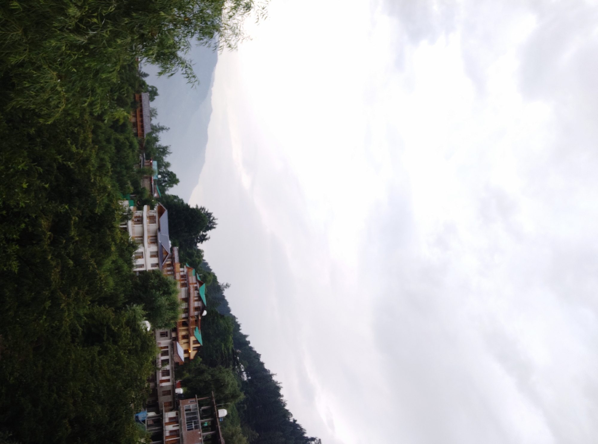 The image depicts a hillside village in Patar, India, characterized by a cluster of multi-story buildings nestled amidst lush green trees. The architecture of the buildings appears to be a mix of traditional and modern styles, with some featuring colorful roofs and balconies. The village is situated on a slope, with layers of houses ascending the hillside. In the background, rolling hills and mountains are visible, partially obscured by clouds. The sky is overcast, suggesting a potentially rainy or misty day. The foreground is dominated by dense foliage, with trees framing the view of the village. The overall impression is one of a serene and picturesque mountain settlement.