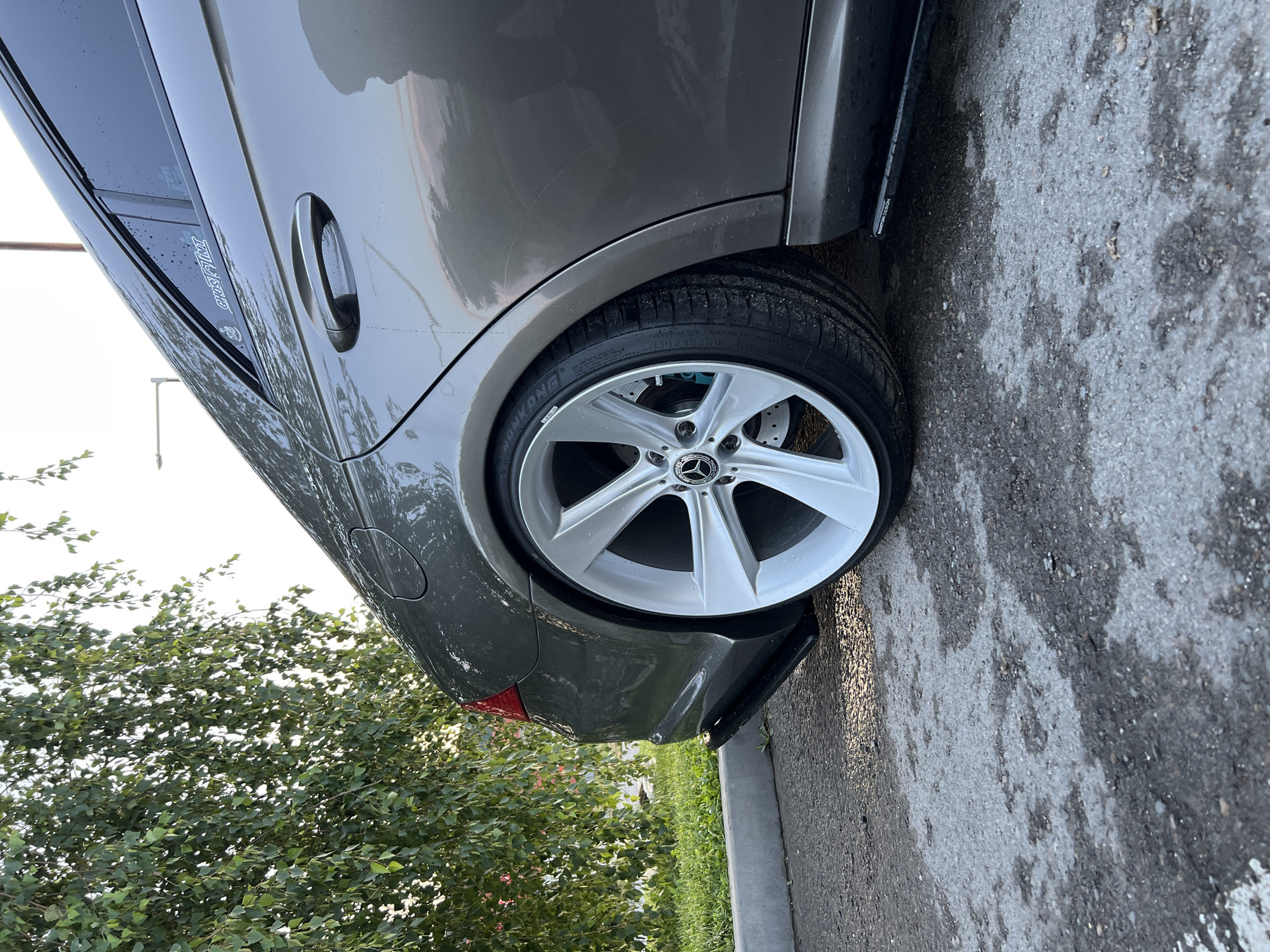 The image, rotated 90 degrees clockwise, depicts the rear passenger side of a dark grey or brown Mercedes-Benz car, appearing to be a sedan or coupe. The vehicle is parked outdoors, likely on an asphalt surface which also shows signs of dampness. The car itself is covered in water droplets, suggesting it has either been recently washed or experienced rain.
The car features prominent silver multi-spoke alloy wheels with a Mercedes-Benz star logo on the center cap. A bright blue brake caliper is visible behind the wheel's spokes. The tire sidewall clearly displays the brand "NANKANG" and the size "235/35ZR19 91Y". Several light scratches are visible on the car's body panel above the rear wheel arch.
On the rear passenger window, a dark deflector or visor is installed, bearing stylized white text that reads "MILOTIRO". The door handle on the rear passenger door is a chrome or polished metallic finish. A part of the red taillight is visible on the far left. A black, possibly aerodynamic, element or side skirt runs along the bottom of the car, displaying small white text that appears to be "M-SPORT DESIGN". A fuel cap is also visible on the rear quarter panel.
In the background, a substantial amount of green foliage from trees and bushes is visible against a bright, clear or overcast sky, indicating a daytime setting.