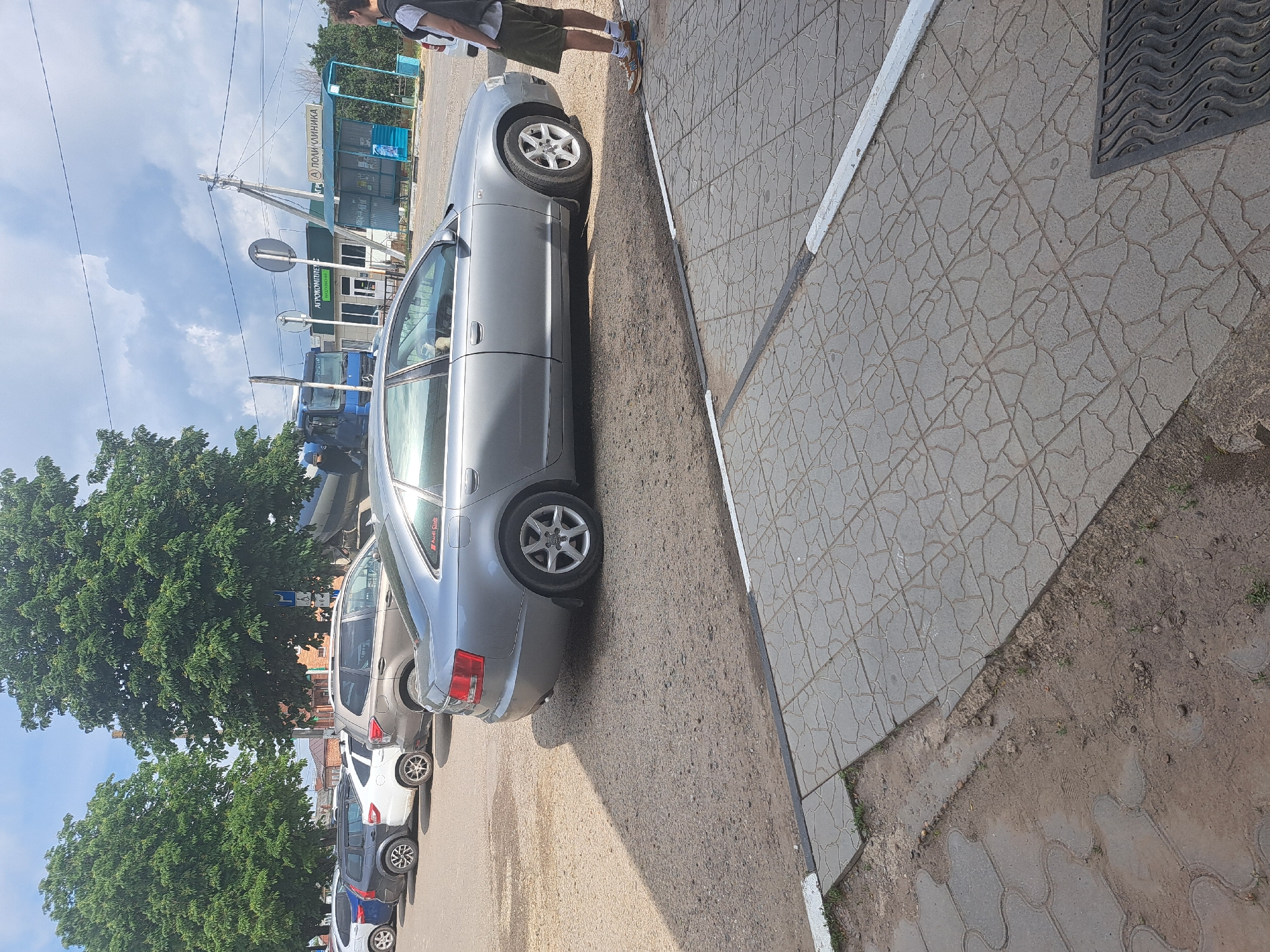 The image depicts a street scene in what is asserted to be Leningradskaya, Russia. The focus is on a light-colored sedan parked along a curb constructed of gray tiles. The vehicle is positioned next to the sidewalk with the passenger side of the car facing the curb. A person, only their lower body visible, stands on the sidewalk. Other cars, including a minivan and a light colored vehicle, are parked further down the street behind the sedan. Trees are present. A bus stop and what appears to be a medical clinic can be seen in the distance. The sky is visible with patches of clouds.