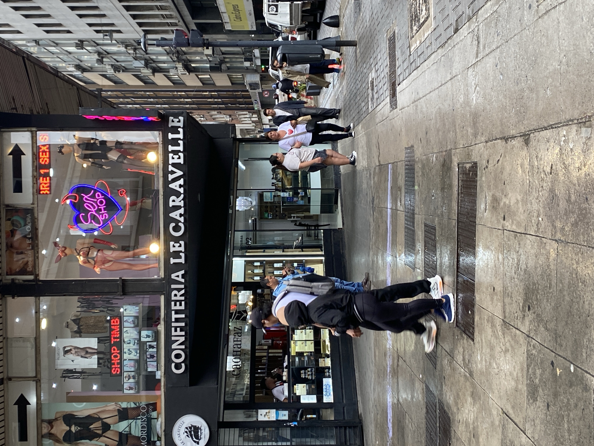 Here is a detailed description of the image:
The image shows a street scene in Buenos Aires, Argentina. On the left side of the frame is a store called "Confiteria Le Caravelle", which also has "Sex Shop" neon sign in one window. The store also has an advertising window displaying lingerie and adult products.
On the sidewalk in front of the store, several people are walking. Towards the bottom of the frame, a person in black pants and white sneakers is walking towards the left. Further up the sidewalk, other people are walking or standing, with one man wearing a white t-shirt with a red heart on it.
The street runs parallel to the storefront, with cars parked along the side. A light-colored truck with "Cobro Express" on it is visible further up the street. Buildings line the street on both sides, creating an urban environment.