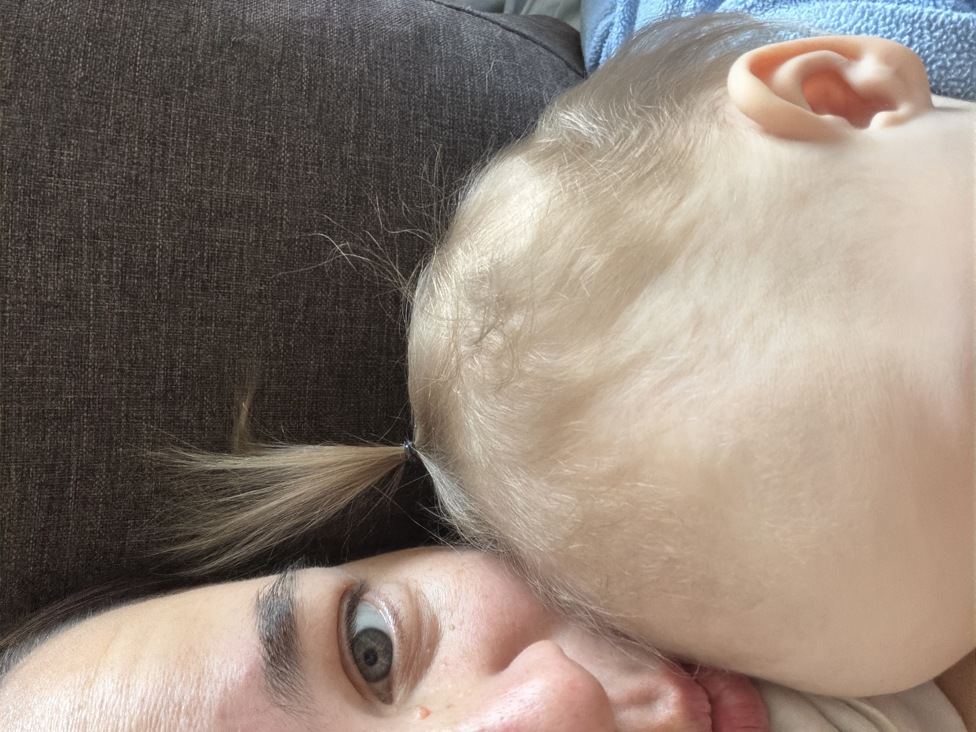 This close-up, portrait-oriented image captures an intimate moment between an adult and a baby or toddler, set indoors in Chaikovsky, Russia. The shot is taken from the adult's perspective, likely a selfie.
On the left side of the frame, the adult's face is partially visible, featuring a wide, expressive grey eye looking directly at the camera, a dark, well-defined eyebrow, and a portion of the nose and mouth area, which is slightly pursed. Small freckles are visible on the adult's skin.
The right side of the image is dominated by the head of a baby or toddler, resting against the adult. The child has very light, fine hair, possibly blonde, with some strands tied into a small, wispy ponytail secured by a tiny dark hair tie. One of the baby's ears, with its distinct curves, is clearly visible. The baby's skin appears fair and smooth.
The background on the left consists of a dark brown or grey textured fabric, likely upholstery from a couch or bed. In the upper right corner, behind the baby's head, a light blue textured fabric, possibly a blanket or towel, is discernible. The lighting is soft and even, suggesting an indoor setting with diffused light.
The activity portrays a moment of close physical bonding, possibly cuddling or resting together. The adult's gaze is directed towards the camera, while the baby's face is obscured by its head resting on the adult, conveying a sense of comfort and affection. No specific time of day, weather conditions, or visible text can be determined from the image.