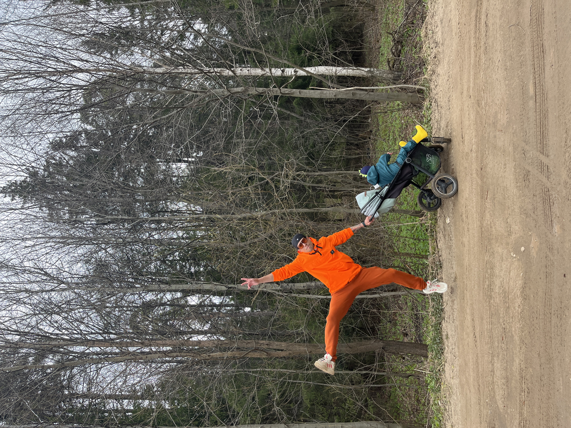 The image, rotated 90 degrees counter-clockwise, depicts an outdoor scene in Chaikovsky, Russia, featuring a man, a child in a stroller, and a natural setting with a dirt path and forest.
In the foreground, a man in vibrant orange clothing performs a playful pose, balancing on one leg with arms outstretched and his other leg extended behind him, as if jumping or taking flight. He wears an orange long-sleeved top, matching orange pants, white and red sneakers, a dark baseball cap, and sunglasses. His left hand forms a 'peace' sign.
To the man's right and slightly behind him, a child is seated in a dark-colored stroller designed for rough terrain. The child is seen from the back, wearing a blue or teal jacket, a dark hat with a blue and yellow pattern (possibly dinosaur spikes), and distinct bright yellow rain boots. Several bags, including a light blue one, are attached to the stroller. A stylized green logo, resembling a leaf or abstract 'B', is visible on the side of the stroller.
The setting is a wooded area with a wide dirt path running along the right side of the frame, showing tire tracks. The background is dominated by a dense forest composed primarily of deciduous trees with bare, intricate branches, suggesting late winter or early spring. Some evergreen trees with green foliage are also visible further back among the bare trees. The sky is overcast, indicating daylight. The overall impression is one of an active and joyful outing in nature. There is no discernible text beyond the described logo on the stroller.