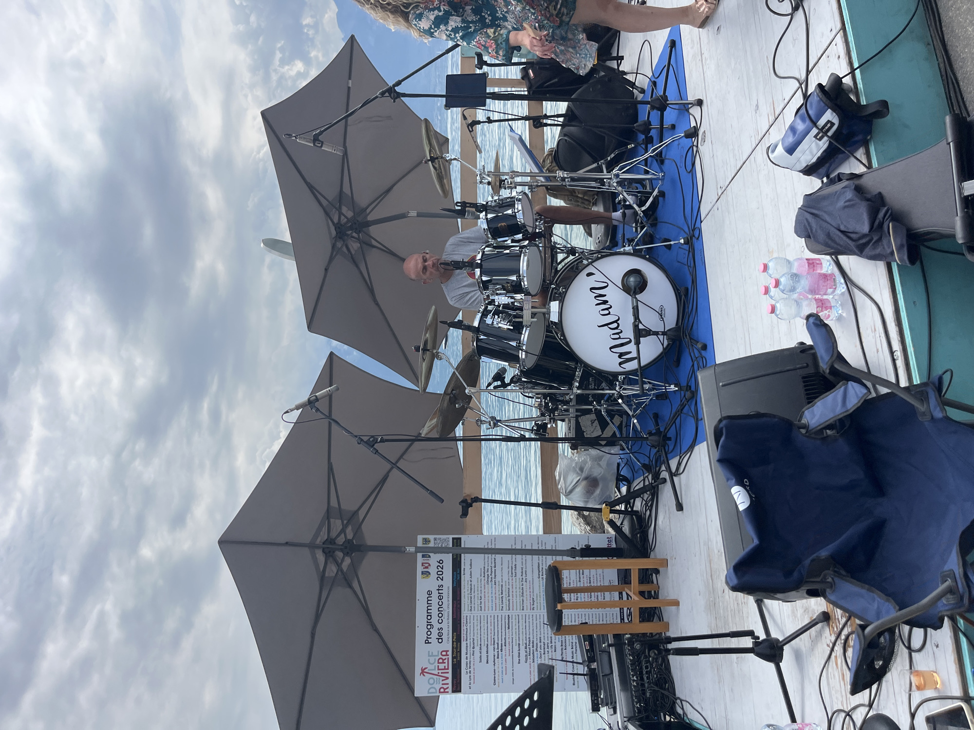 An outdoor concert stage is set up on a wooden deck overlooking a body of water, likely Lake Geneva, in Vevey, Switzerland. A male drummer, wearing a grey t-shirt and shorts, is seated behind a drum kit. The drum kit has a large bass drum with the word "Madam" written on it. Two large grey umbrellas provide shade over the drum set and the surrounding area. A poster board listing "Programme des concerts 2026" is visible to the left of the drummer. Various musical equipment, including microphones, stands, speakers, and cables, are scattered across the stage. A blue folding chair is positioned in the foreground. A woman, partially visible on the right side of the image, is wearing a floral dress and sandals. The sky is overcast with clouds. The scene appears to be a performance or preparation for a performance at the Riviera.