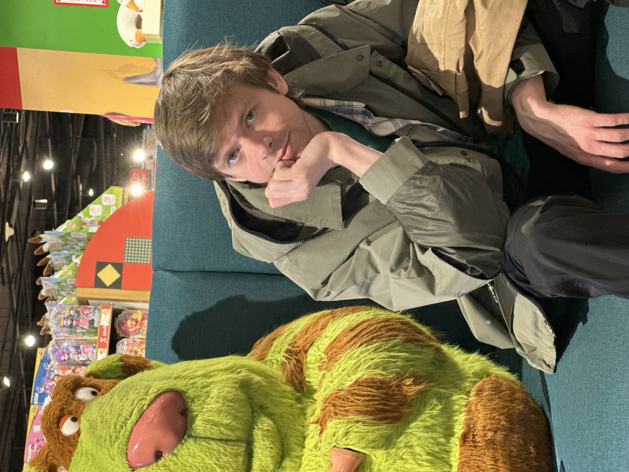 The image shows a man reclining on a blue couch next to a large, plush toy dog. The man has light brown hair and is wearing a green jacket over a plaid shirt. He rests his chin on his hand, looking towards the camera. The dog toy is mostly green with brown stripes. The background shows a store aisle with toys and displays. Based on the store's branding, the location is Detsky Mir (Children's World) in Moscow, Russia.