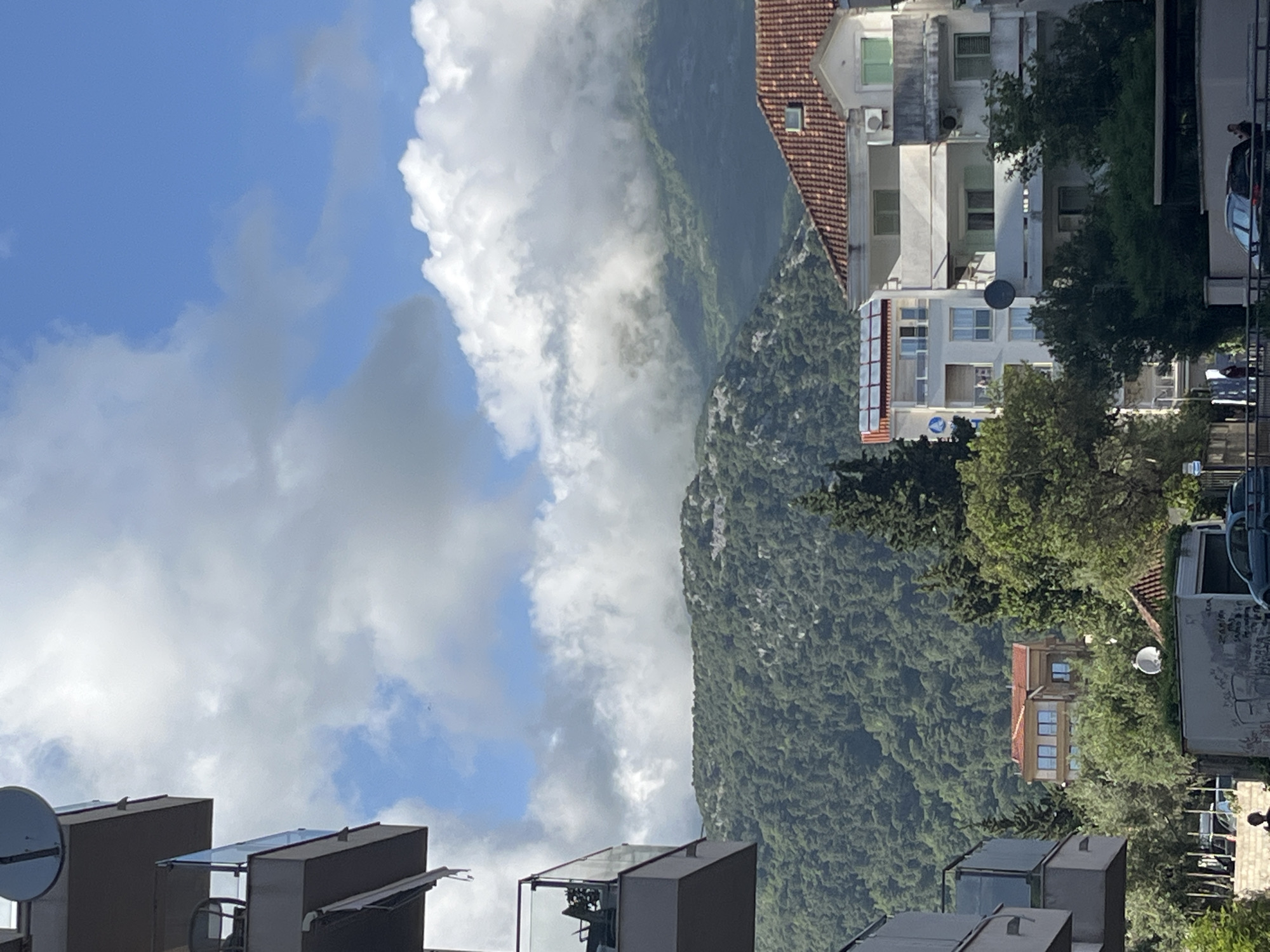 The image captures an outdoor scene in Budva, Montenegro. A densely forested mountain rises in the background, partially obscured by white, puffy clouds. In the foreground, there are rooftops of buildings with modern, boxy structures. A building with a red tile roof and white walls is seen mid-ground. The sky is blue with scattered clouds. Trees and other vegetation are also visible, growing around the buildings and on the mountain. Cars and satellite dishes can be identified on some buildings.