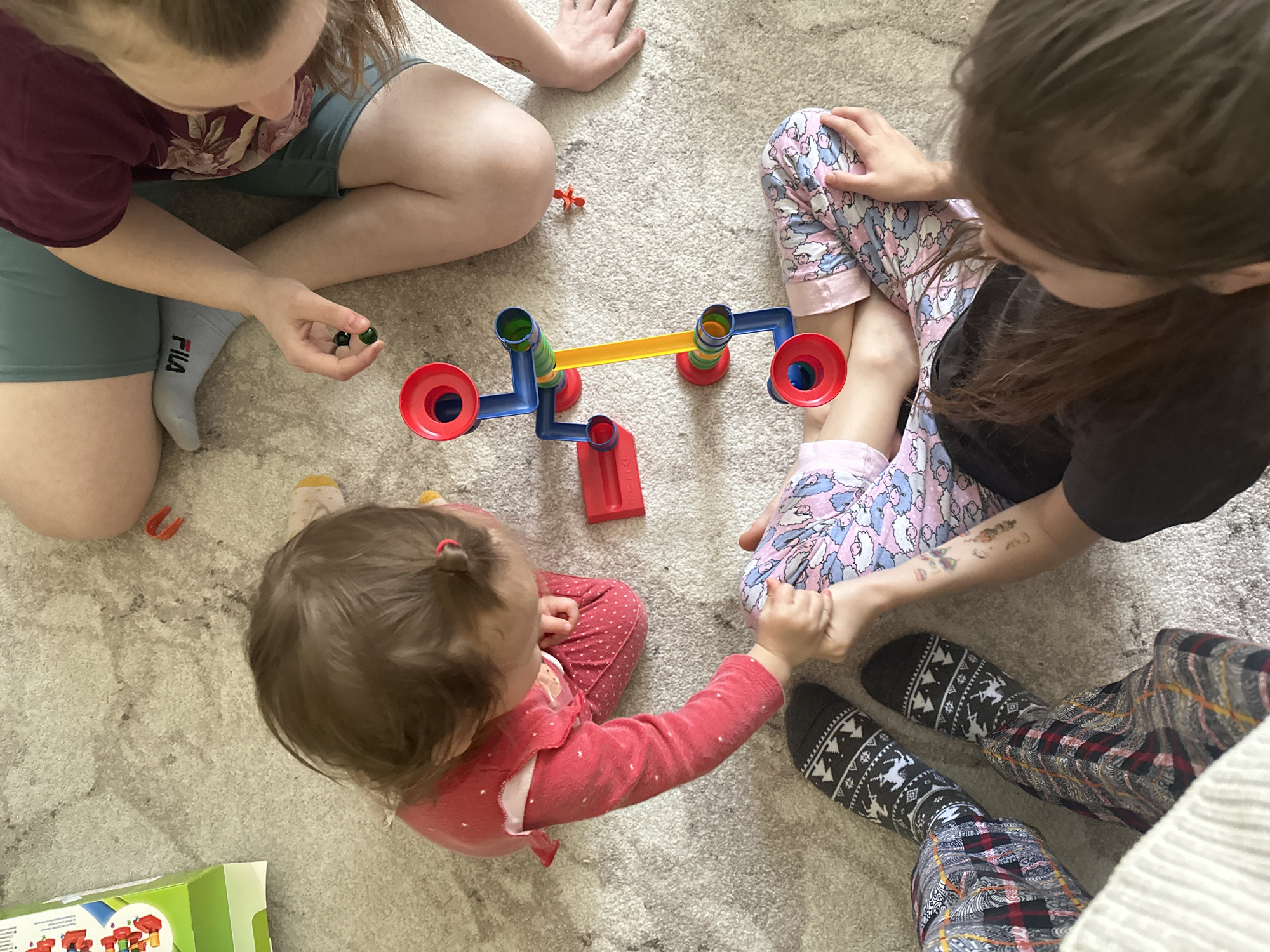An overhead shot captures a group of people gathered on a light-colored, patterned carpet, engaged in playing with a colorful balancing toy. The scene is set indoors, likely in a home in Uva, Russia.
In the upper left, an individual wearing a maroon floral t-shirt, light blue shorts, and a white sock with "FILA" visible, holds a dark green marble, seemingly poised to place it on the toy. To their right, another person is seated, identifiable by pink and white patterned pajama pants featuring sheep or clouds, a black t-shirt, and dark patterned socks with geometric and reindeer designs. A tattoo is visible on their lower leg.
At the bottom center, a young child or toddler, with a small ponytail secured by a red hair tie, wears a red polka-dot long-sleeved top. They are looking towards the toy and reaching out, showing interest in the activity. The lower legs and patterned socks of a third individual are visible in the bottom right, suggesting multiple people are involved or observing the play.
The central object is a bright, multi-colored balancing toy, possibly a marble run or scale. It features blue structural components, red funnel-like cups on either end, a yellow connecting beam, and green and blue vertical supports. A small orange plastic piece lies on the carpet near the top-left person's knee. Part of a green and white box, likely the toy's packaging, is visible in the bottom left corner. The atmosphere appears casual and focused, with everyone engaged in the play.
