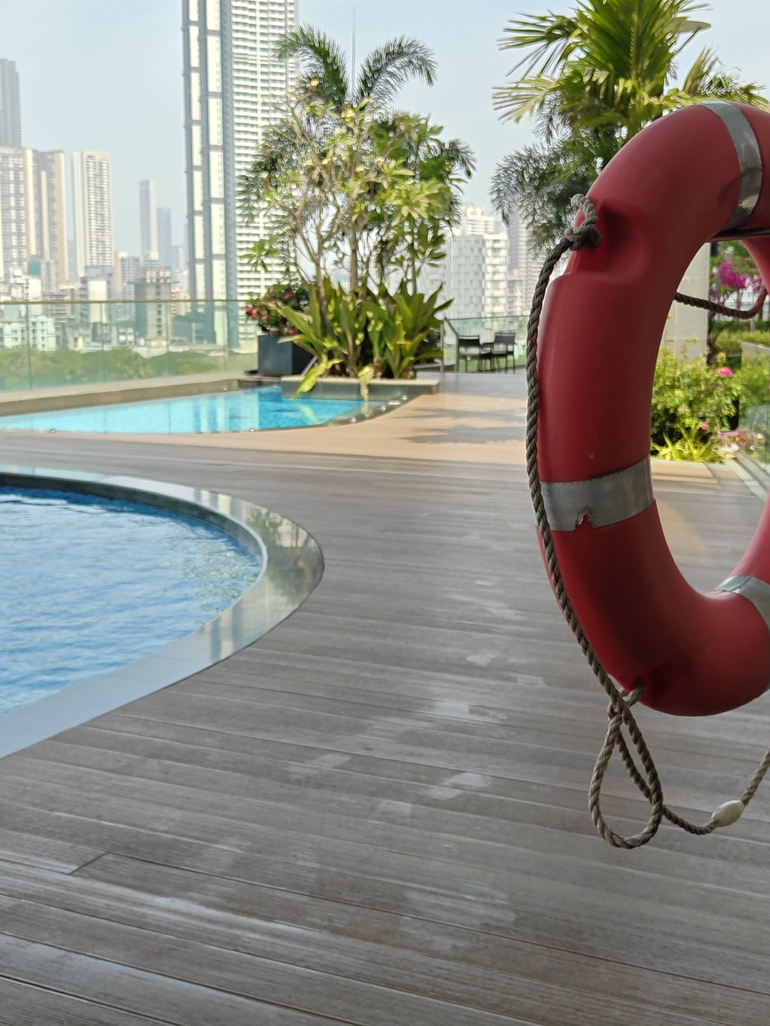 This image captures a bright, daytime scene of a luxurious rooftop pool area, offering a panoramic view of a bustling city skyline, specifically Mumbai, India.
In the foreground and prominently featured on the right side is a vibrant red lifebuoy (life preserver) with a white rope looped around it. The lifebuoy has two reflective silver bands, one of which shows some wear. The rope, made of braided natural fiber, hangs down from the lifebuoy, forming a loose knot.
To the left of the lifebuoy, two swimming pools are visible. The closest pool, partially seen in the lower left, is circular with clear blue water and a shiny silver-colored edge. Further back, another rectangular pool also features inviting blue water, reflecting the clear sky. Both pools are surrounded by a clean, light brown wooden deck, which extends throughout the visible ground area. The wooden planks show some water marks or weathering in places.
Beyond the pools and deck, lush green foliage, including several palm trees and potted plants with some pink flowers, adds a touch of nature to the urban environment. A transparent glass barrier separates the pool area from the expansive cityscape. In the mid-ground, a few outdoor chairs and a table can be seen, suggesting a seating area.
The background is dominated by numerous high-rise buildings and skyscrapers under a bright, somewhat hazy sky, characteristic of a large metropolitan area like Mumbai. The distant buildings appear in various shades of grey and white, receding into the haze.
No people are visible in the image, contributing to a serene and tranquil atmosphere. There is no visible text in the scene. The lighting suggests it is either morning or late afternoon.