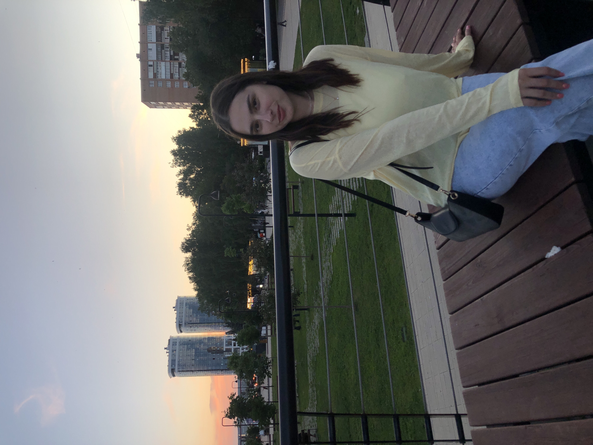 A medium shot captures a young woman with long, dark hair and fair skin sitting on a wooden bench in a park. She is wearing a light yellow, long-sleeved shirt and light blue jeans. A black crossbody bag is slung over her shoulder. Her hands rest on the bench, and her fingernails are painted a bright orange. The background shows a green grassy area with paved pathways, trees, and a black metal railing. In the distance, two modern high-rise buildings with reflective glass facades stand against a soft, orange and blue twilight sky, characteristic of a sunset. A more traditional brick apartment building is also visible. The scene appears to be in a public park within the city of Izhevsk, Russia.
