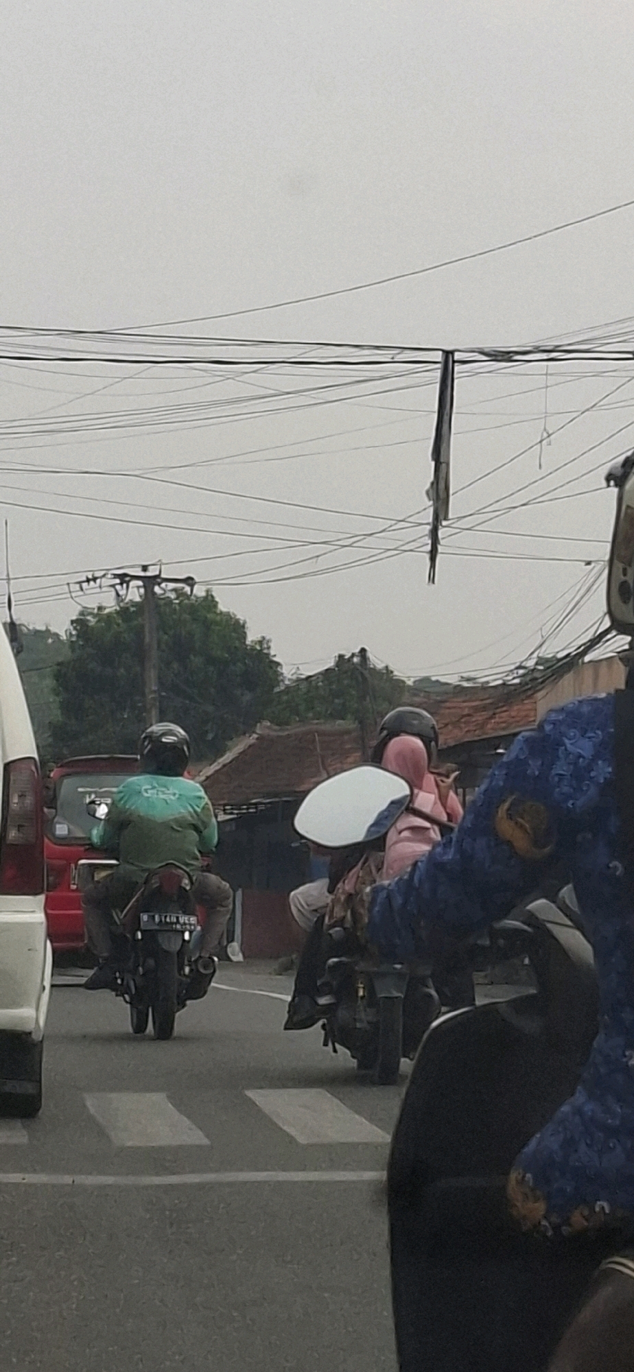 The image depicts a street scene in Sirnagalih, Indonesia, under an overcast sky. In the foreground, a person on a motorcycle, wearing a blue jacket with embroidered detailing and a helmet, is partially visible. Behind them, another motorcycle carries two passengers, one in a pink hijab and the other in a green jacket with a "Grab" logo. The passenger in the pink hijab is looking back. To the left, the rear of a white vehicle is visible, and further back, a red vehicle. The background shows a street with electrical wires overhead, a utility pole, and houses with tiled roofs, suggesting a residential or mixed-use urban area. The road surface is asphalt with white markings, possibly indicating a pedestrian crossing.