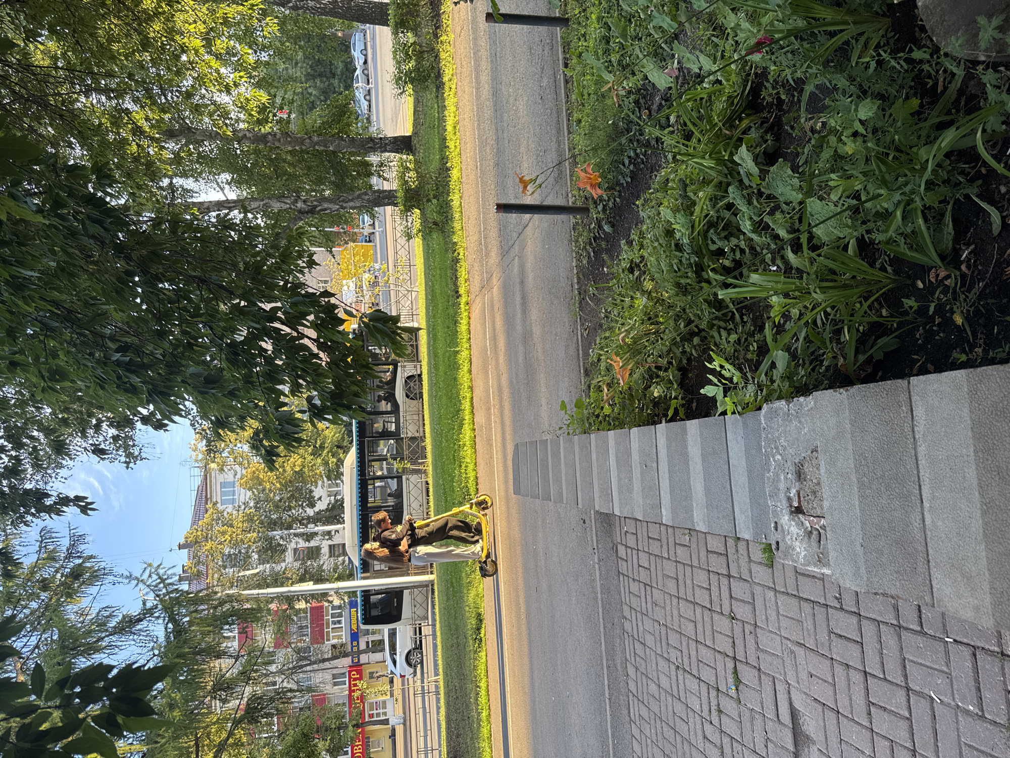 The image shows two people on a yellow electric scooter, stopped on a paved path next to a road. The man is standing and appears to be adjusting something on the scooter or the woman, who is seated. The woman is looking at him. They are dressed casually.
The scene is an urban environment with trees lining the street and buildings in the background. A bus is visible on the road, and a white car is parked further down. The path has a brick pattern on one side and a concrete curb with some vegetation growing over it on the other. There are also some orange flowers growing in the green area beside the path. The sun is casting long shadows, indicating it is likely late afternoon or early morning. The sky is blue with some wispy clouds.
Visible text on the bus includes Cyrillic letters, and there are signs on the buildings in the background also in Cyrillic. The overall mood seems relaxed and casual.