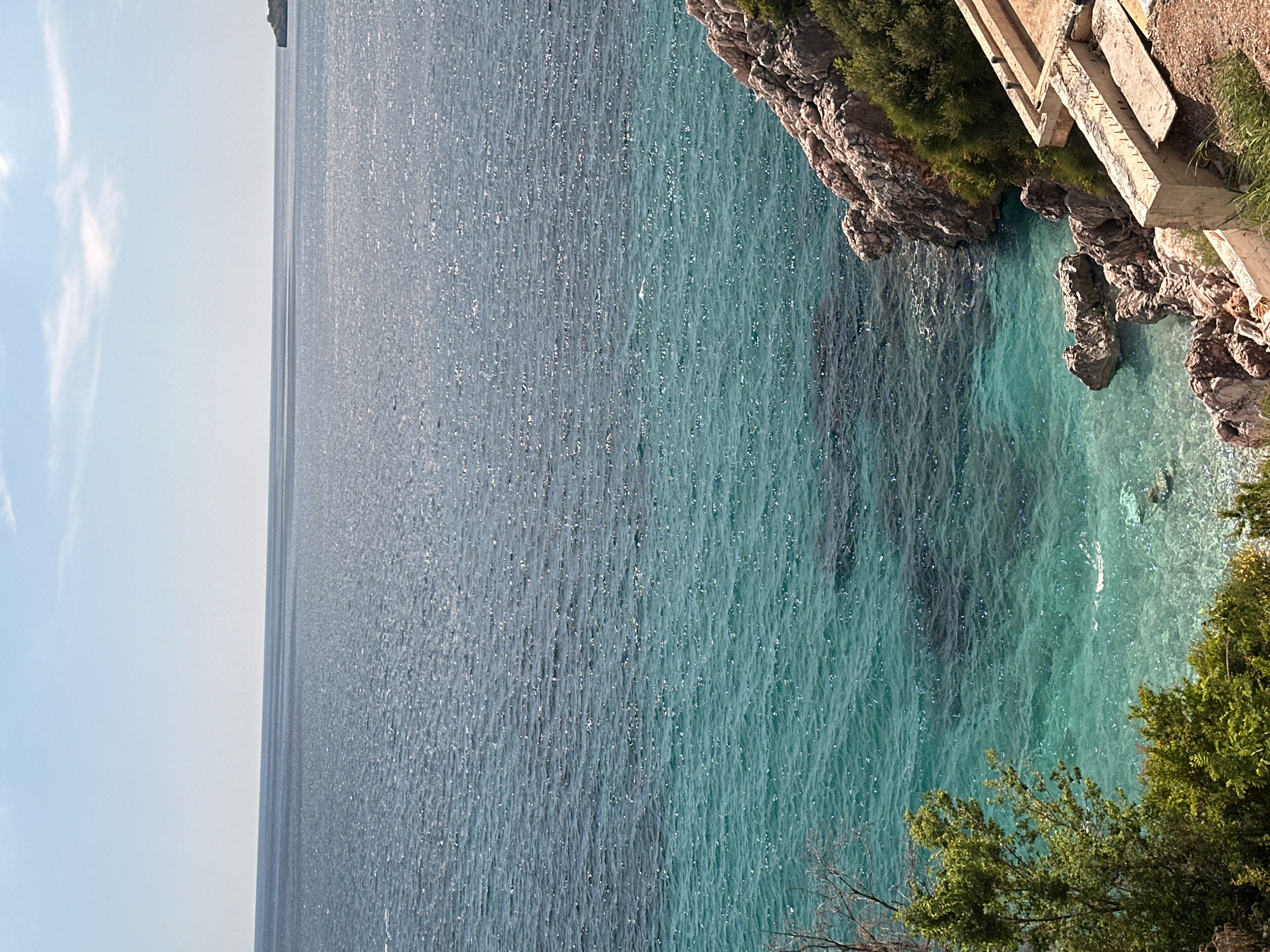 This media file captures a vibrant coastal landscape in Kamenovo, Montenegro, presented with a vertical orientation (originally rotated 90 degrees counter-clockwise). The scene is dominated by the sparkling sea, with a clear blue sky and a rocky coastline.
The upper left portion of the image shows a bright, clear blue sky with a few wispy white clouds, indicating sunny weather. The vast expanse of the sea stretches across the majority of the frame, from the horizon to the foreground. The water exhibits two distinct shades: a darker, deeper blue in the open sea towards the left, and a brilliant, clear turquoise closer to the coastline on the right. Both sections of the water surface are covered in countless shimmering reflections from the sun, suggesting small ripples or waves.
On the right side, a rugged coastline composed of rocks and cliffs meets the sea. Patches of lush green vegetation, including trees and bushes, grow along the cliff edges and in the foreground at the bottom right. Nestled into the cliff face on the upper right, there is a man-made concrete structure, possibly a set of stairs or a pathway, appearing somewhat weathered. Faint, dark markings or graffiti are visible on its surface, but they are illegible.
There are no people or specific activities taking place; the image depicts a serene natural landscape. The bright sunlight, clear sky, and vibrant water colors suggest it is daytime, likely morning or early afternoon. The location cues, such as the clear turquoise water, rocky shore, and Mediterranean-style vegetation, are consistent with the provided context of Kamenovo, Montenegro.