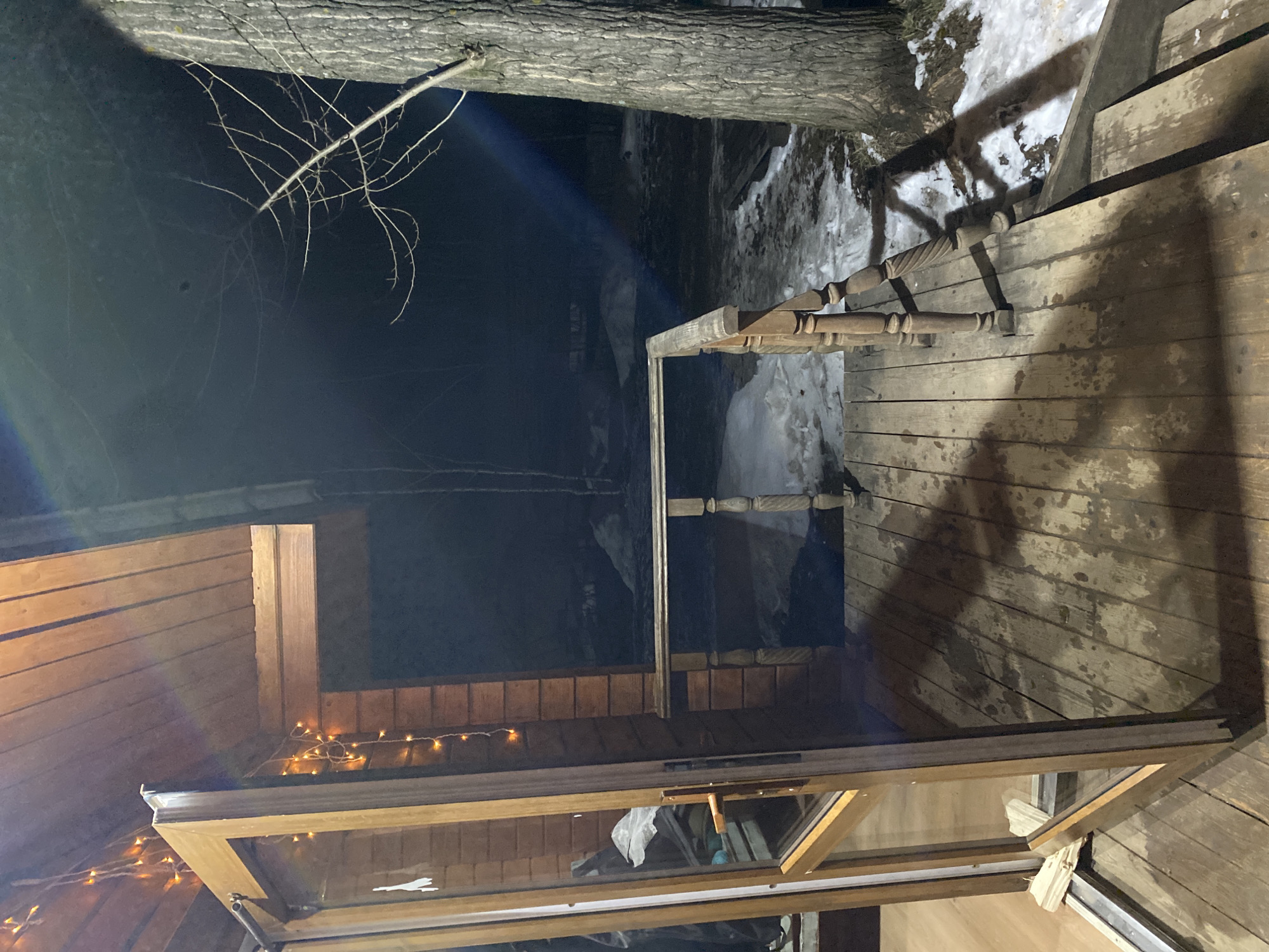 The image shows a night scene in Chaikovsky, Russia. A wooden porch is visible, with wooden planks and a railing. The ground outside the porch is covered in snow and ice. A tree trunk is visible near the edge of the porch. An open window with a wooden frame is present, adorned with string lights. The darkness suggests a rural or sparsely lit area.