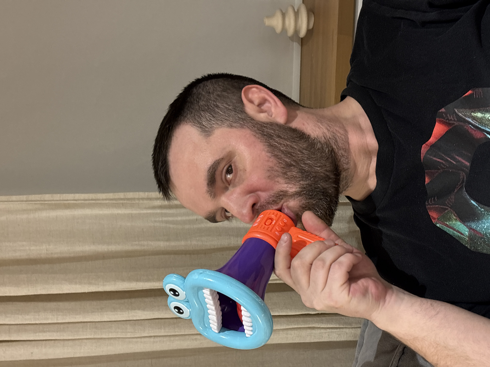 A man with a beard is holding a toy monster megaphone to his mouth. The megaphone is blue with cartoon eyes and white teeth. The middle section is purple and the handle is orange. The man is wearing a black t-shirt and his hair is dark and trimmed close. The background is a pale gray wall with beige horizontal stripes. A white decorative fixture is attached to a wooden door frame in the upper right. The scene appears to be indoors. There is no information to indicate a location in Chaikovsky, Russia.