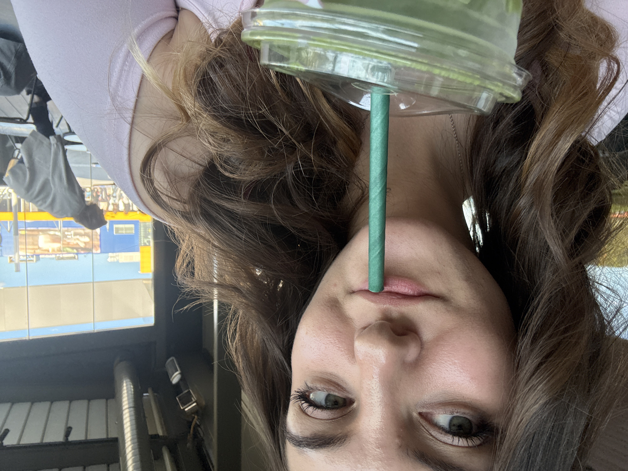 An eye-level portrait shows a young woman, appearing to have light-toned skin, with her head tilted slightly. Her long, wavy brown hair cascades around her shoulders. She wears a light-colored top, possibly pink or lavender. Her eyes, which appear to be a green or hazel color, are looking slightly upwards or directly at the camera. A green paper straw extends from a clear plastic cup into her mouth. The cup, partially visible at the top of the frame, contains a green beverage. A thin, metallic chain necklace is visible around her neck.
The background is out of focus but provides contextual clues. It suggests an indoor or semi-outdoor commercial setting, possibly a cafe or food court, featuring large windows or glass panels. Reflections in these panels show urban buildings with blue and yellow elements under a bright, clear sky, indicating daytime. Interior details in the blurred background include what appear to be industrial-style ceiling fixtures, such as silver-colored ductwork and lighting. The woman's expression is neutral and calm. There is no clearly legible text visible in the image. The scene is set in Sofia, Bulgaria.