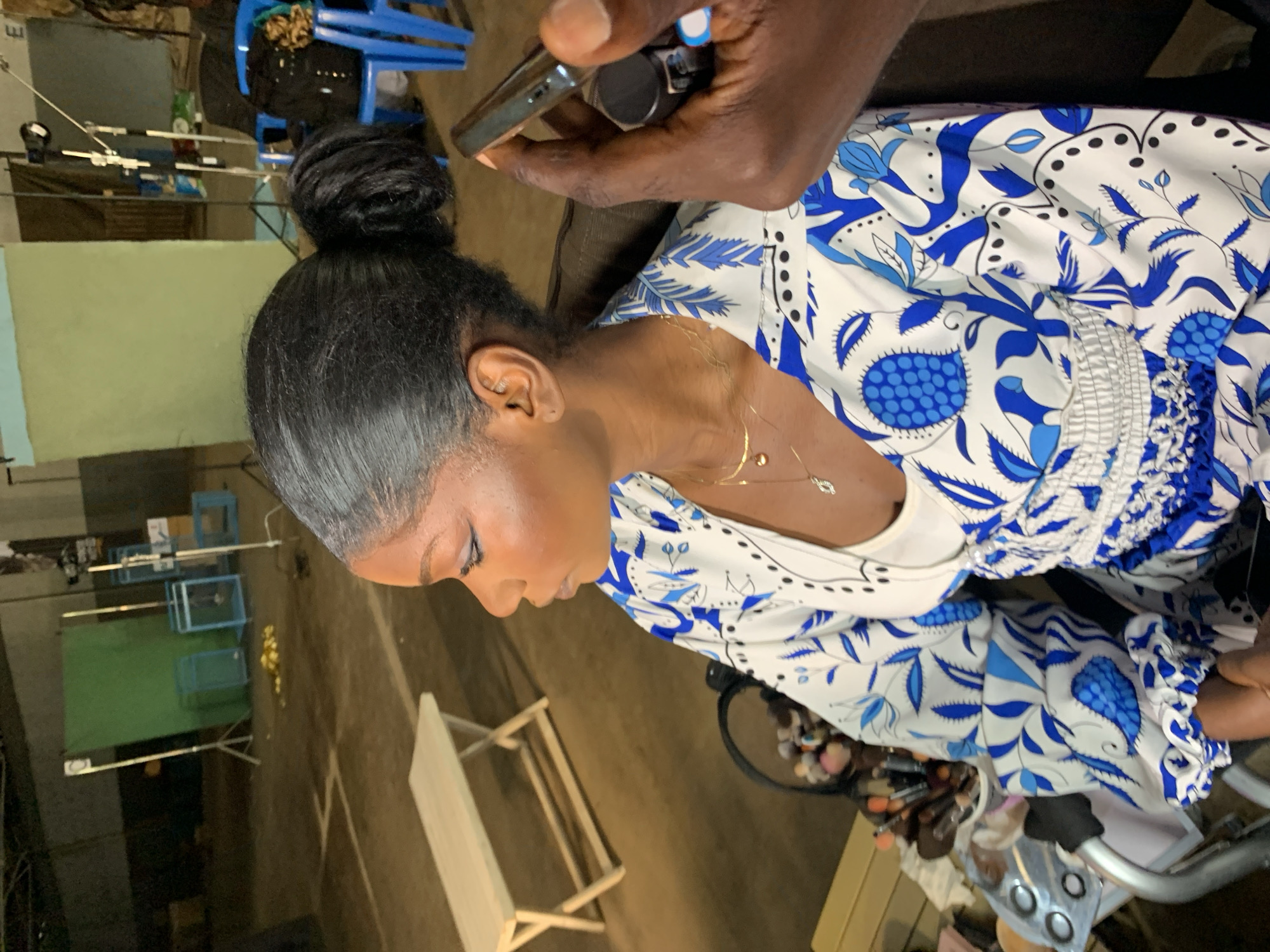 A person is having their makeup done in what appears to be a studio or warehouse setting. The person is wearing a white and blue floral patterned dress. In the background, there are lights, backdrops, and chairs, suggesting a professional setup. A makeup artist's hand is visible holding a phone.