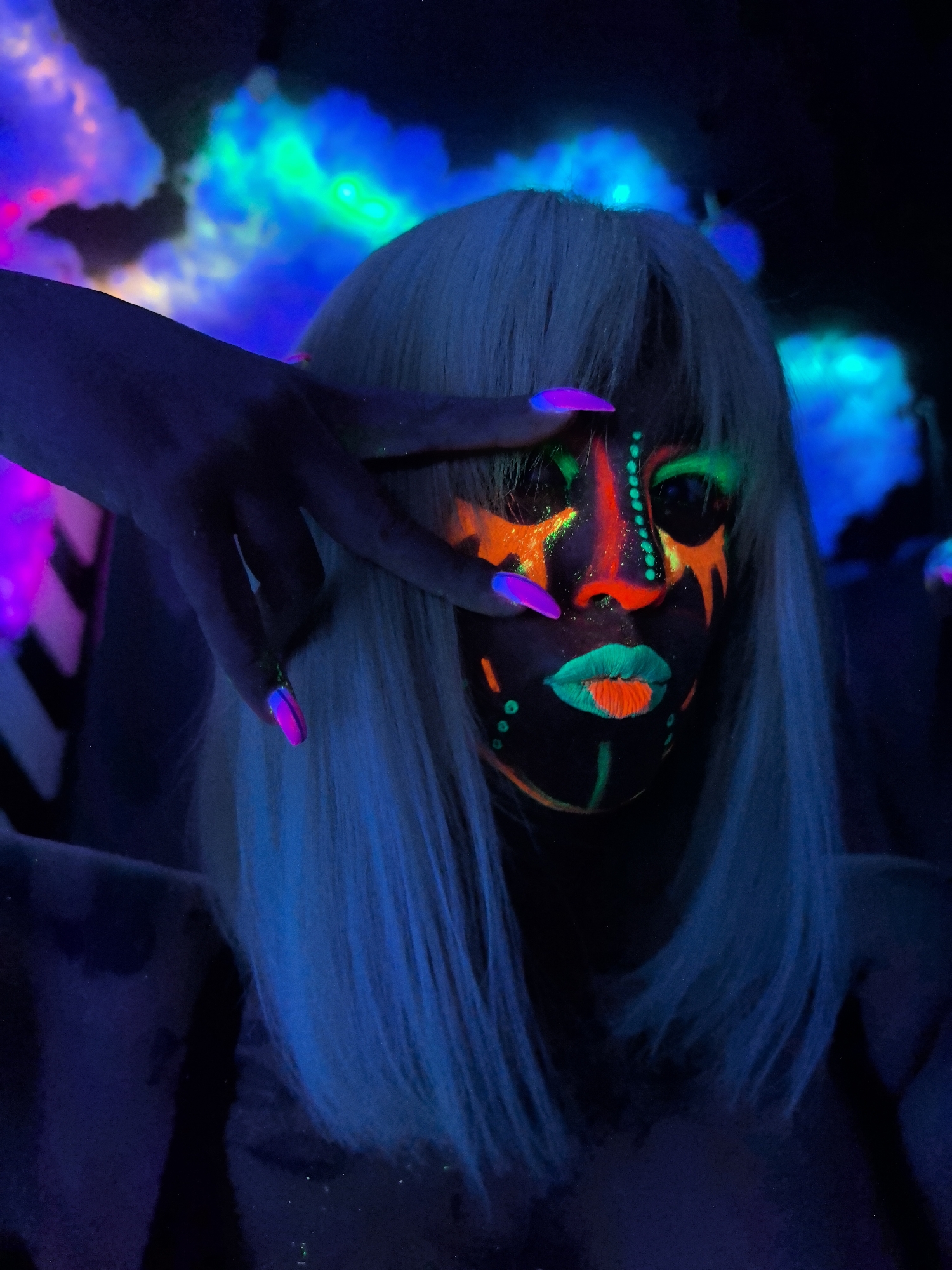 A person is depicted in a medium close-up, illuminated by blacklight in a dark setting. The central subject is a person, likely a woman, with long, straight, light-colored hair that appears white or very light blonde, glowing faintly blue under the ultraviolet light. She has elaborate, vibrant, and intricate face paint in neon orange, bright green, red, and pink, which glows intensely under the blacklight. The makeup includes dots, lines, and abstract shapes, highlighting the nose, mouth, and areas around the eyes. Her lips are painted neon green with an orange stripe, and her nose is bright red-orange. Her eyes are dark, blending into the surrounding blackness.
One hand is raised and partially covers her face, with fingers spread, obscuring the upper left side of her face. Her nails are long, pointed, and feature a striking two-tone effect, glowing bright pink/purple and blue.
The background is dark and out of focus, featuring blurry, luminous formations that resemble smoke or clouds, glowing in various neon colors including blue, green, and purple/pink. This suggests an indoor setting, possibly a club, concert, or artistic performance space, where UV-reactive materials and lighting are intentionally used. The activity appears to be posing for a photograph, showcasing the fluorescent makeup and accessories. The overall mood is artistic, futuristic, and vibrant. There are no specific location cues visible in the image to indicate Noviy, Russia, nor any visible text. The time of day is certainly night or a controlled dark environment. Emotions are conveyed through the bold artistic expression rather than direct facial features, which are largely stylized by the makeup.