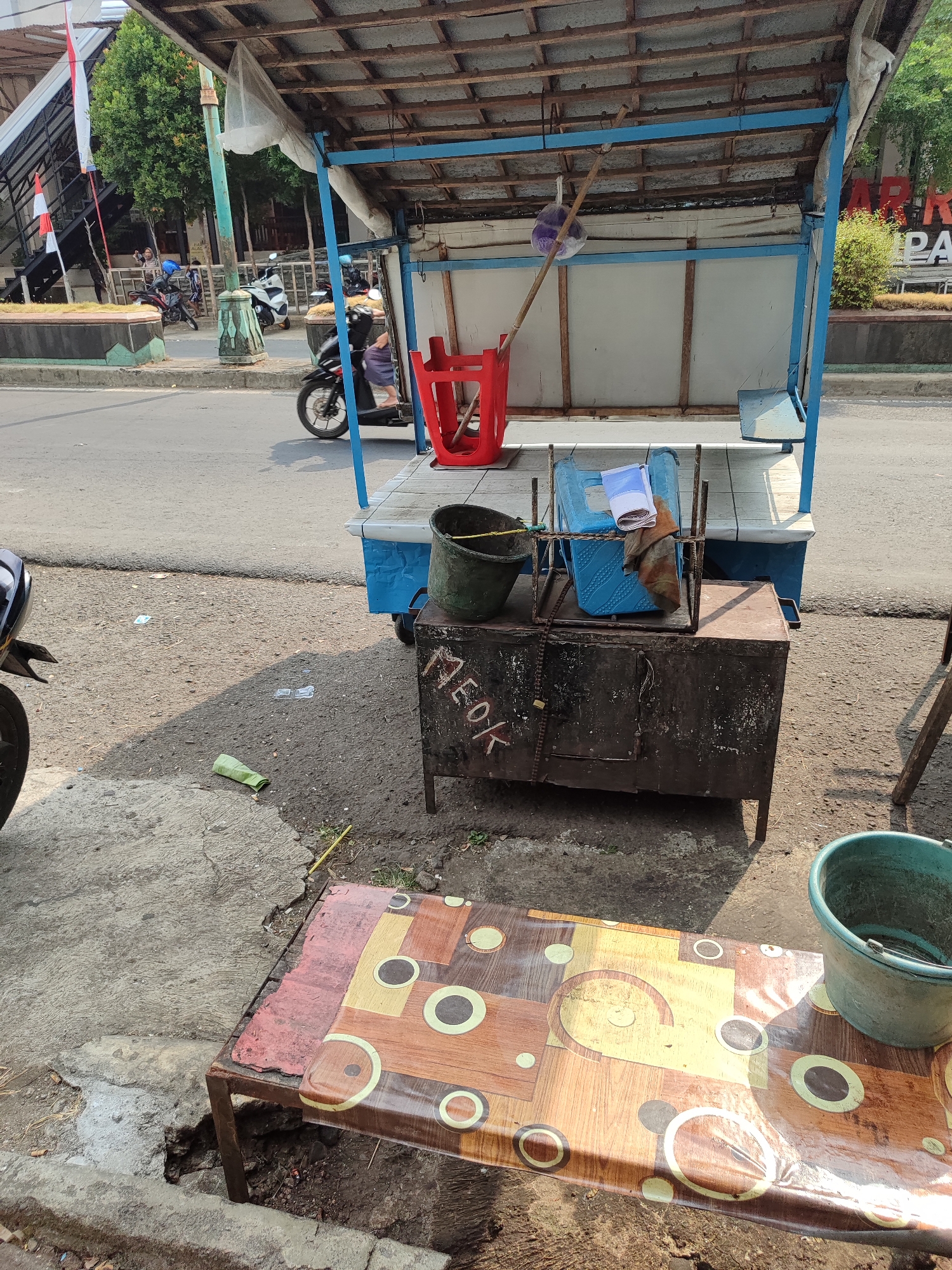 The image depicts a street scene in Randudongkal, Indonesia, likely in front of a small business or street vendor. In the foreground, a portable table with a patterned plastic cover is set up. To the right of the table, a green plastic bucket is visible. Behind the table, a dark, metallic cabinet with legs stands on the ground. The cabinet has some markings, including "MEOK" in white letters. A chain is attached to the cabinet, and a rusty metal structure with blue supports is above it. This structure appears to be part of a mobile cart or stall, with a wooden platform and a roof made of corrugated metal and a white tarp. A red plastic chair is placed on the platform. In the background, a street runs horizontally across the image. Several motorcycles are parked or in motion on the street. In the far background, there is a pedestrian walkway with stairs, decorated with Indonesian flags. Trees and buildings are also visible. The ground is paved with asphalt and concrete, with some debris scattered around. The lighting suggests it is daytime, with shadows cast on the ground.
